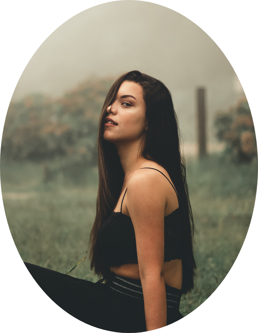 A young woman with long dark hair wearing a black spaghetti strap top and black pants sitting outdoors with a foggy, blurred background of trees and open field.