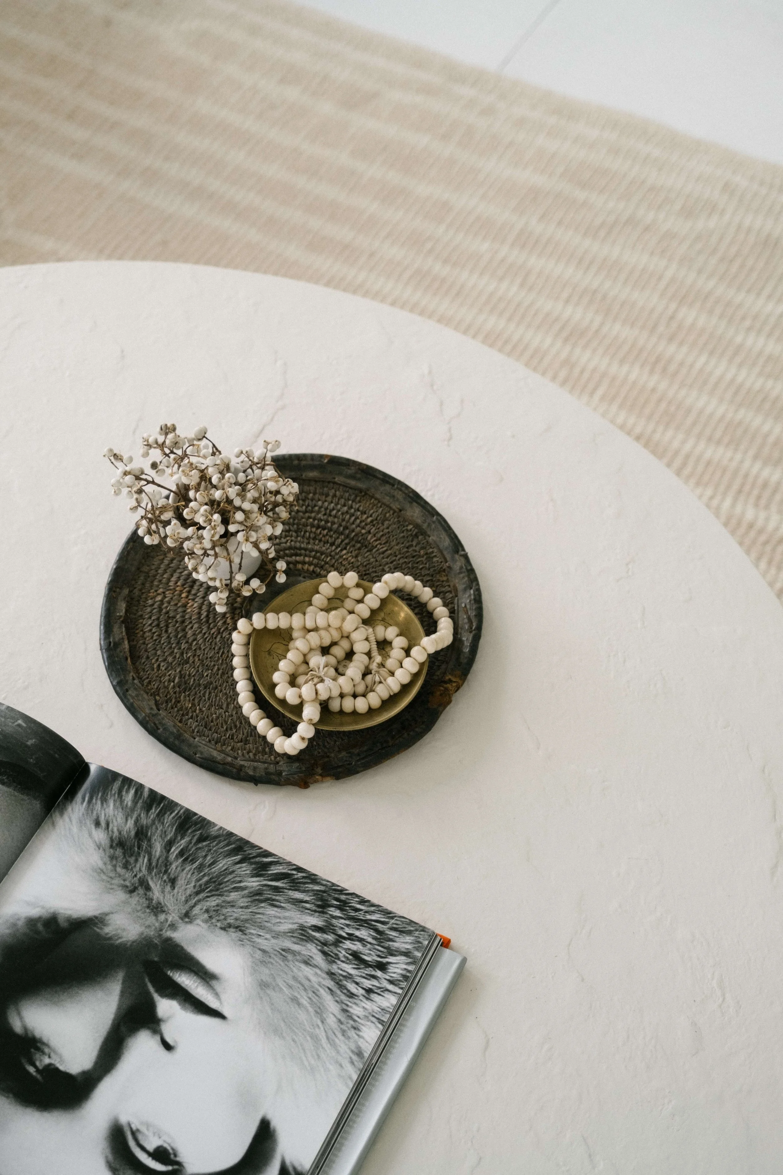A round white table with a small black woven basket containing a sprig of dried white flowers and a beaded necklace. An open magazine with a black-and-white cat's face and eye close-up, resting on a white surface next to the table.