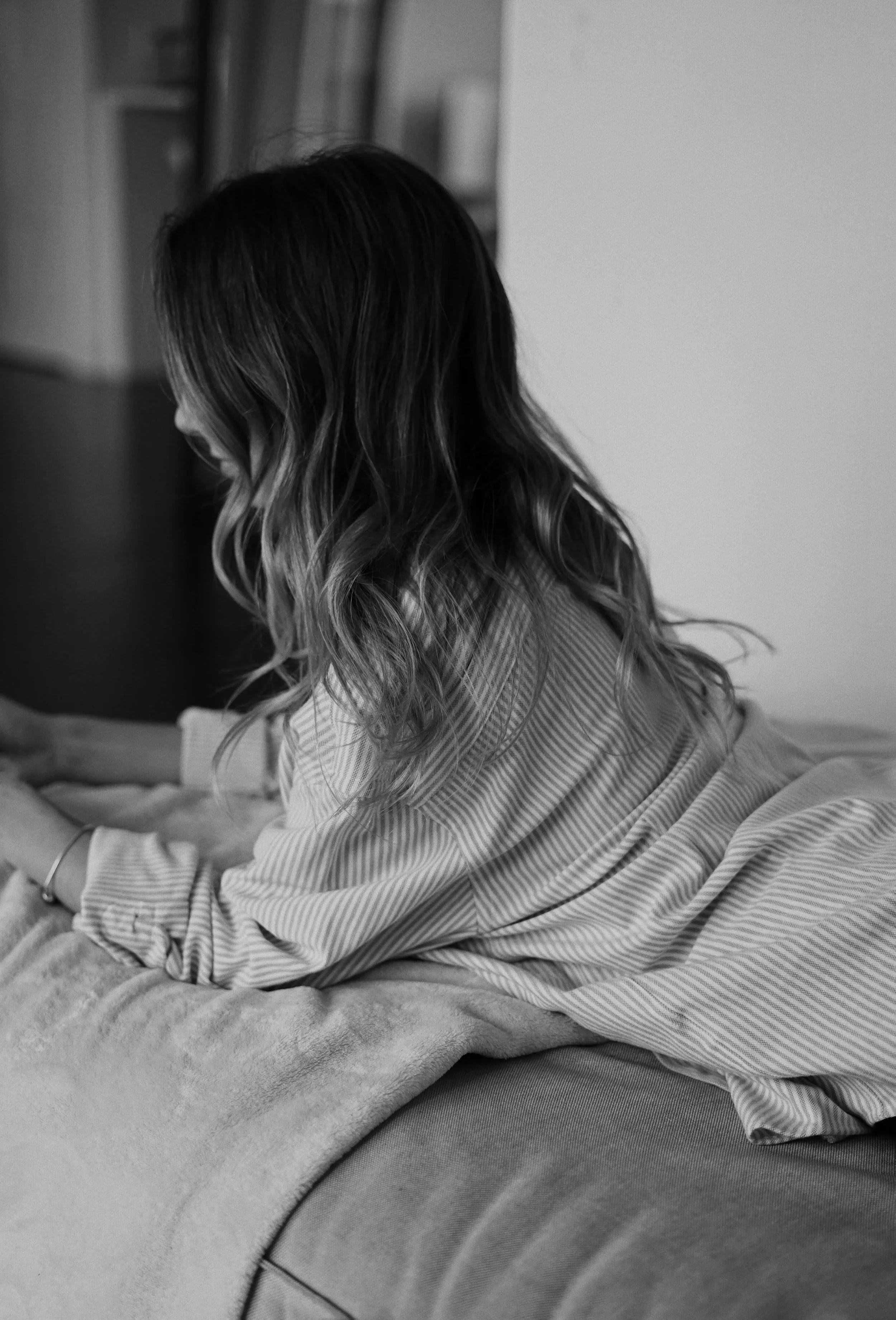A woman with wavy hair sitting on a bed, facing left, wearing a striped shirt and pants, with a blanket or pillow in front of her, in a black and white photo.