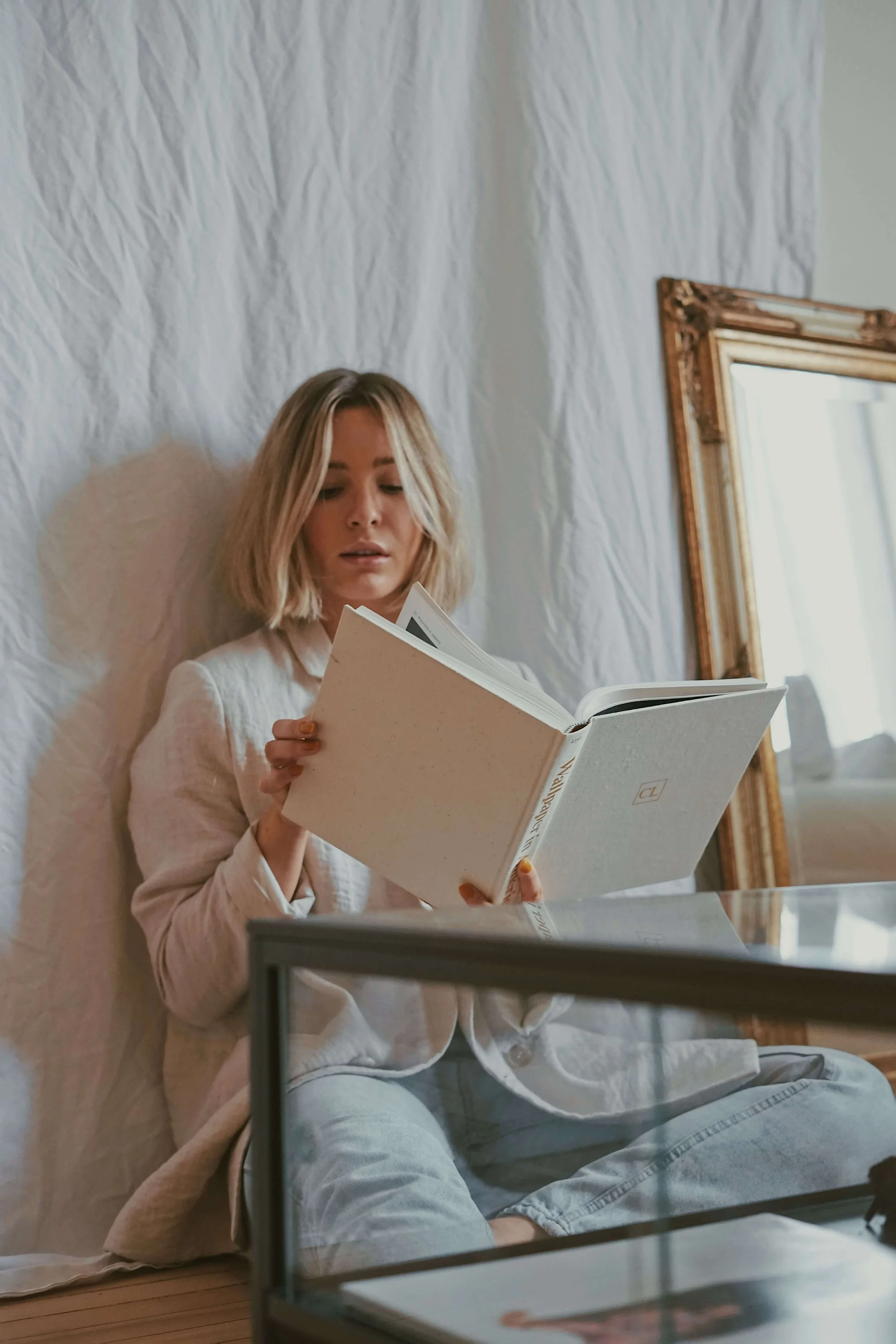 A young woman with blonde hair sitting on a beige cushioned chair, reading a white hardcover book in a room with plain off-white walls, a large mirror with an ornate gold frame, and a glass table in front of her.