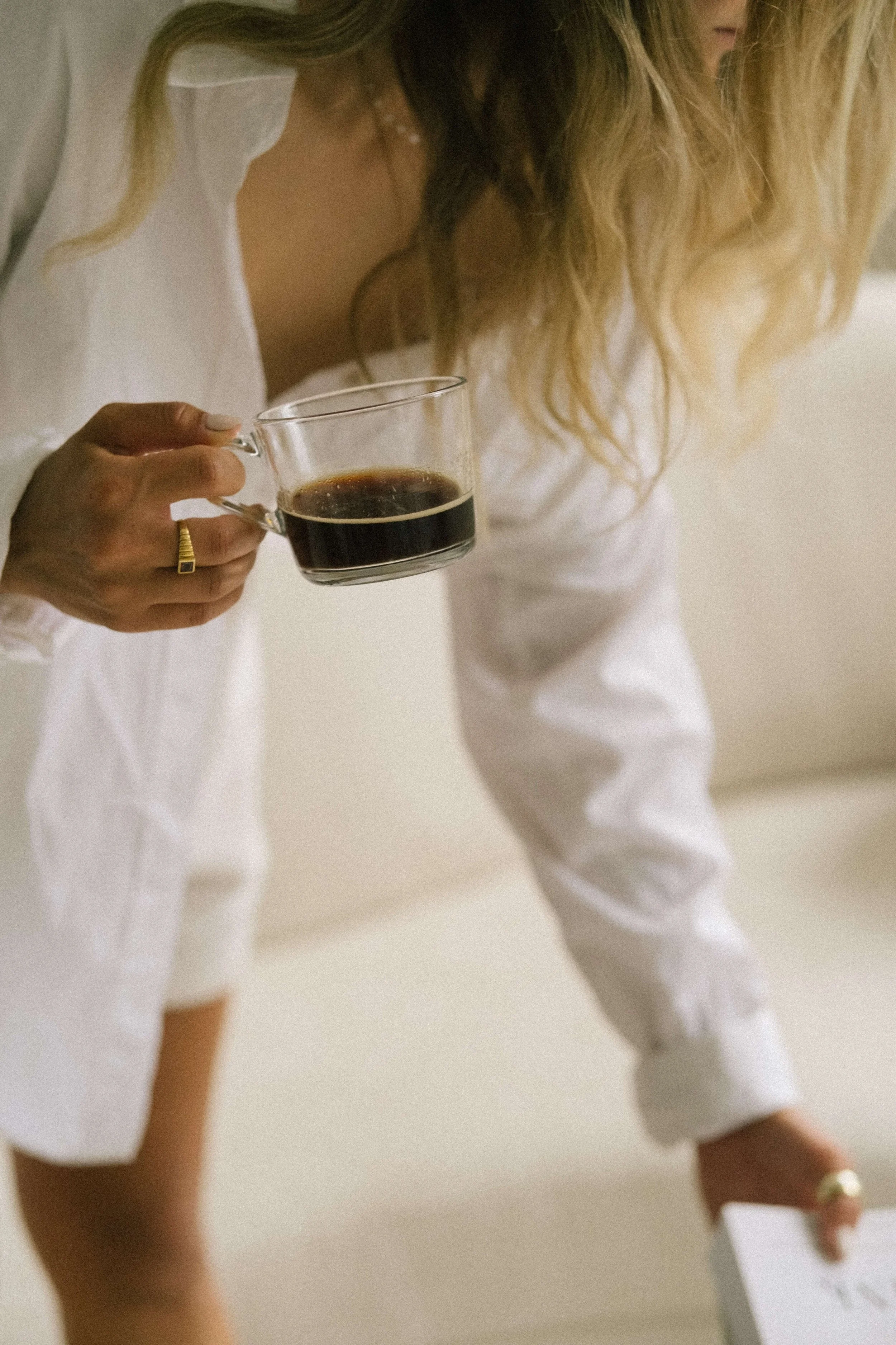 A woman with blonde hair holding a cup of black coffee, wearing a white top and jewelry, with a blurred background.