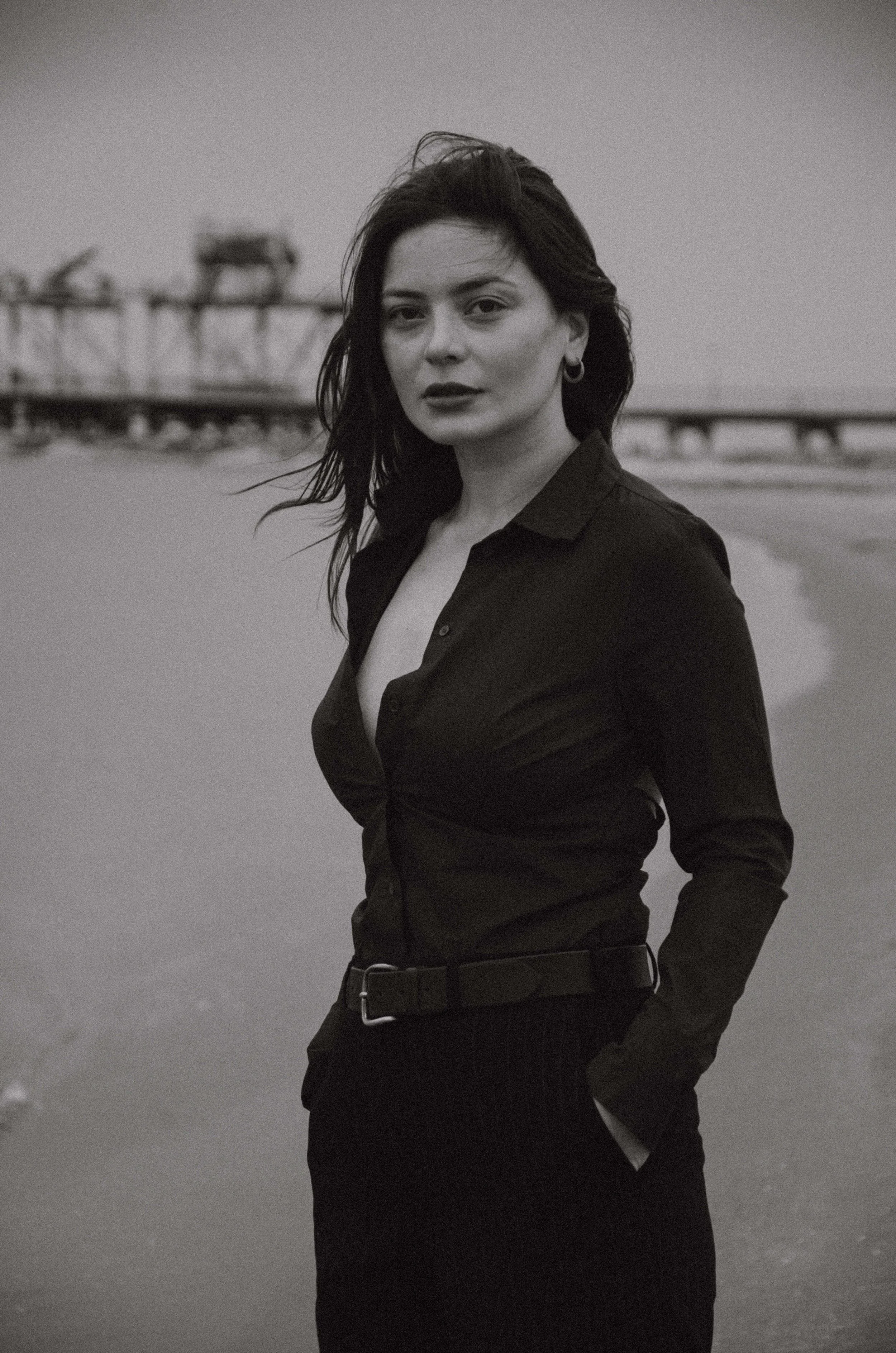 A woman with dark hair wearing a black shirt and pants standing on a beach with a pier in the background, black and white photograph.