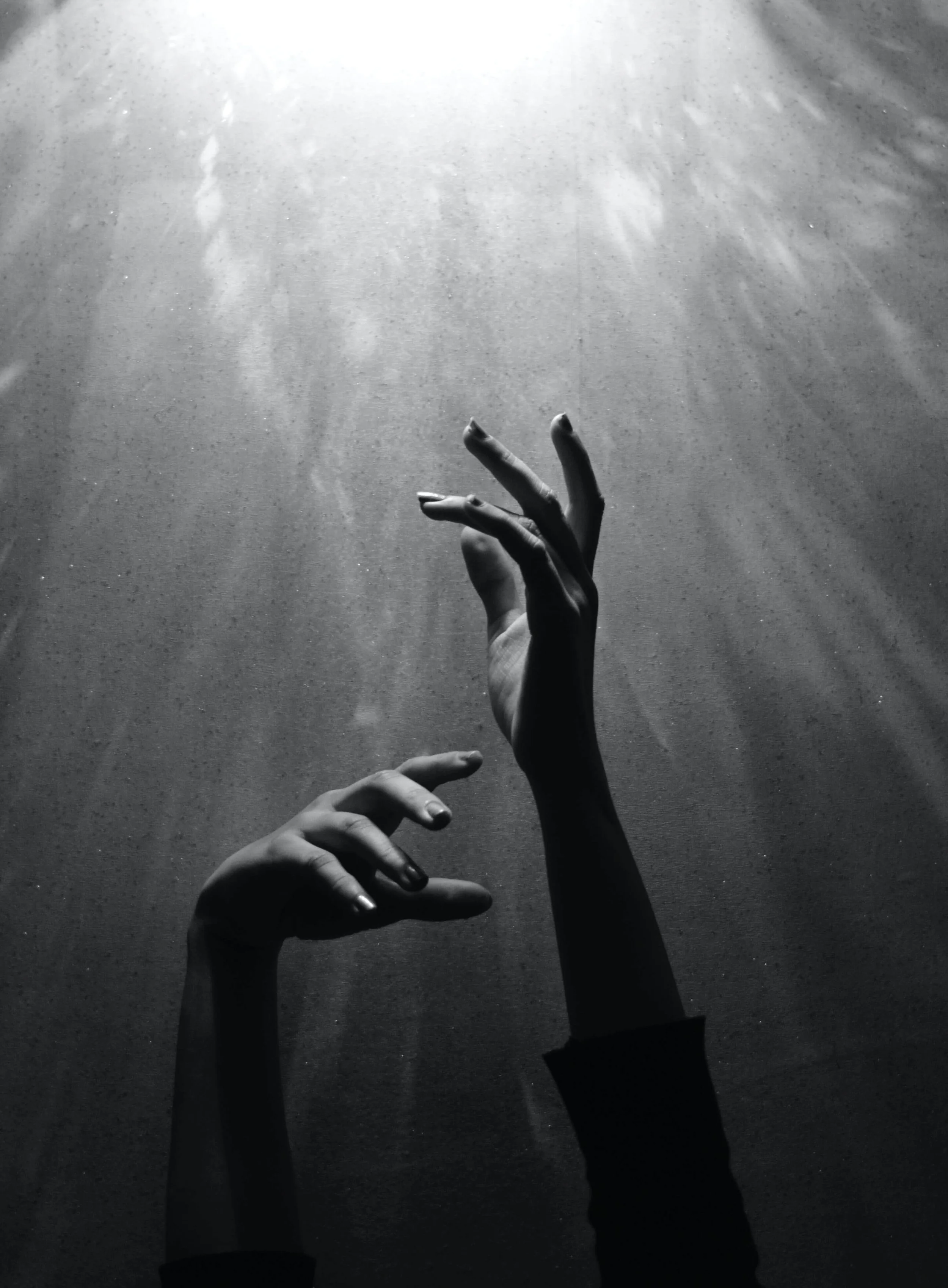 A black and white photo of multiple hands reaching up and outward onto a textured surface, with the light coming from above.