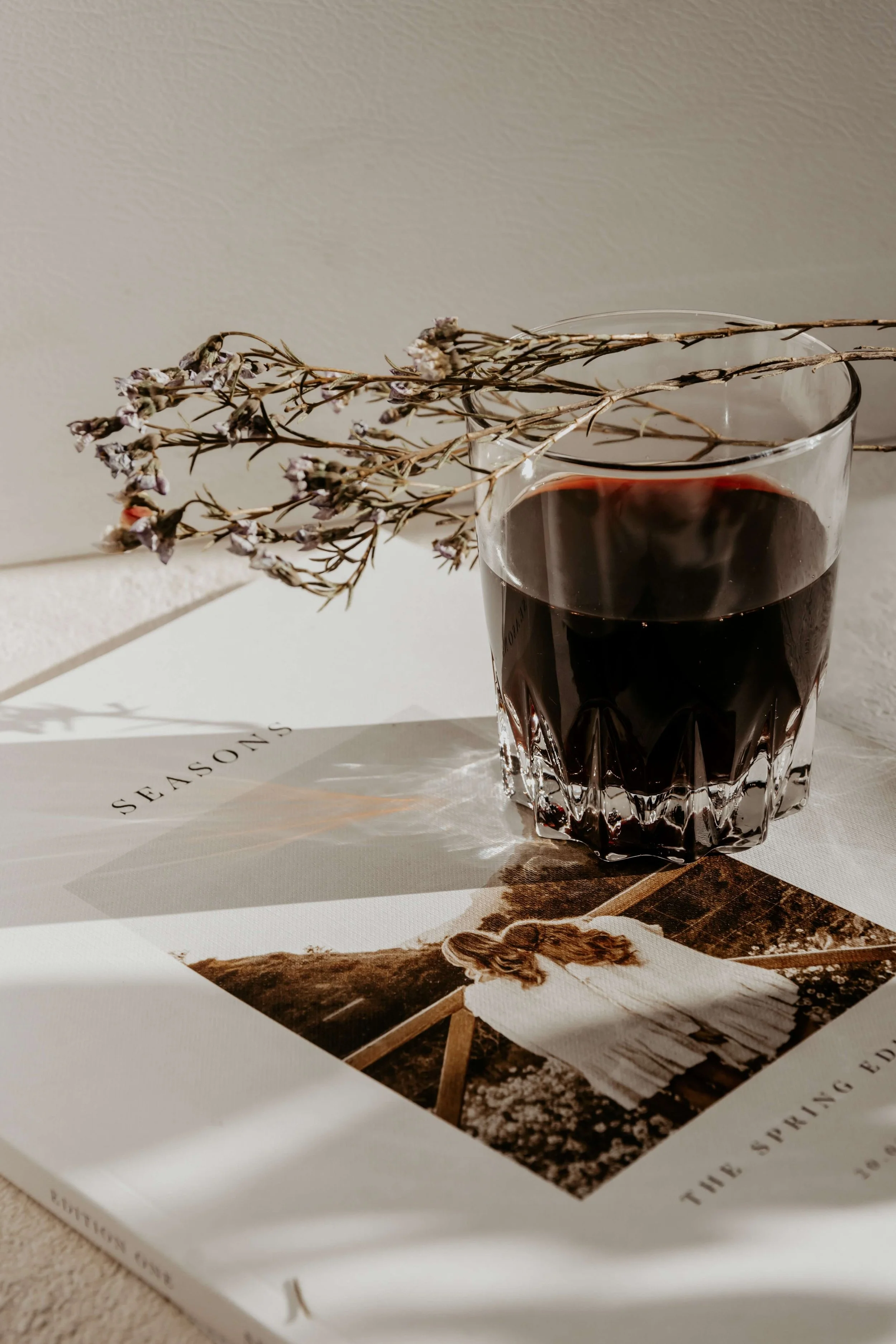 A glass of red wine rests on a magazine with a photograph of a person lying on a bench in a park, and a sprig of dried lavender is placed on top of the glass.