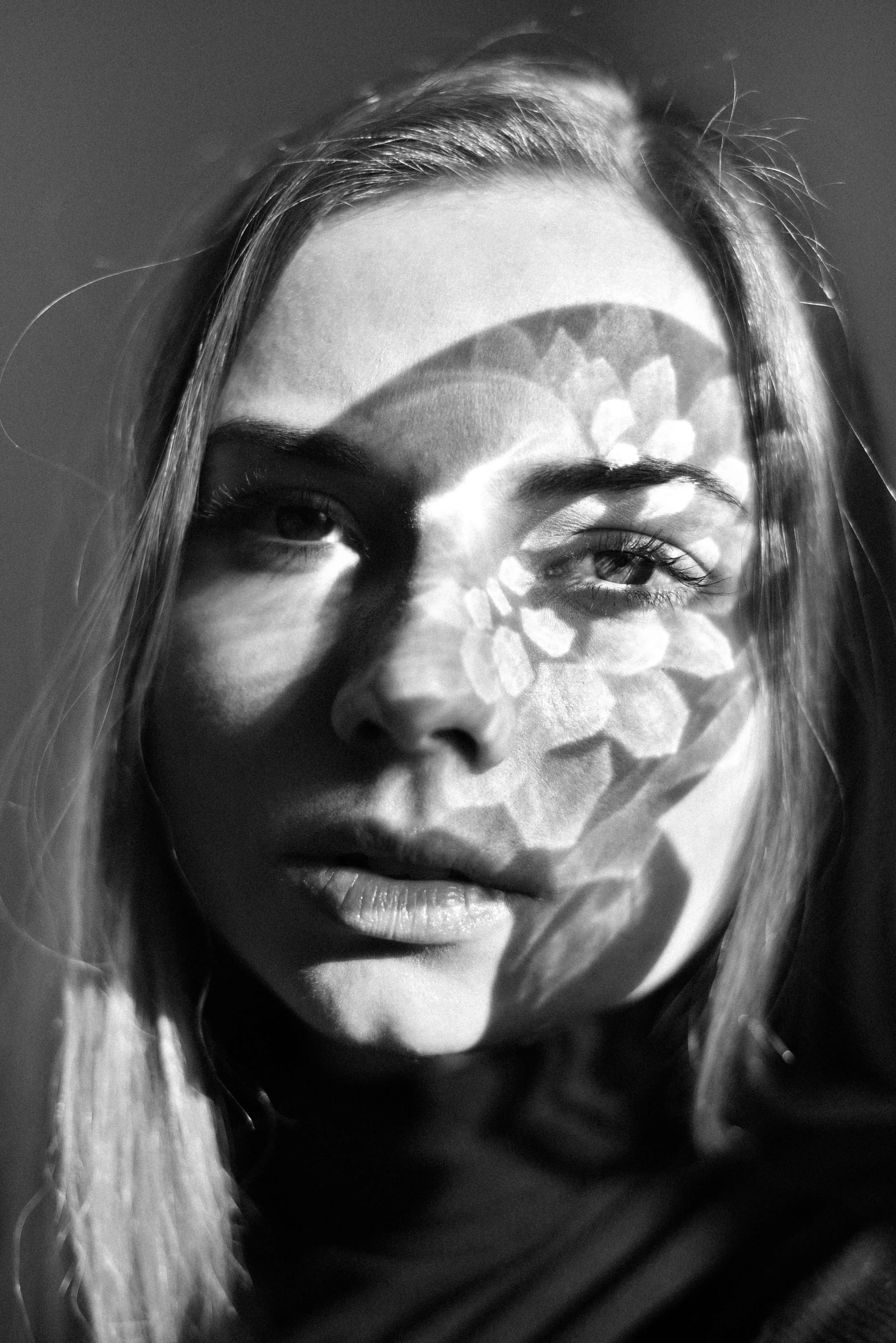 Close-up black and white photo of a woman's face with a patterned shadow cast across her forehead and face, her eye half-closed, and her lips slightly parted.