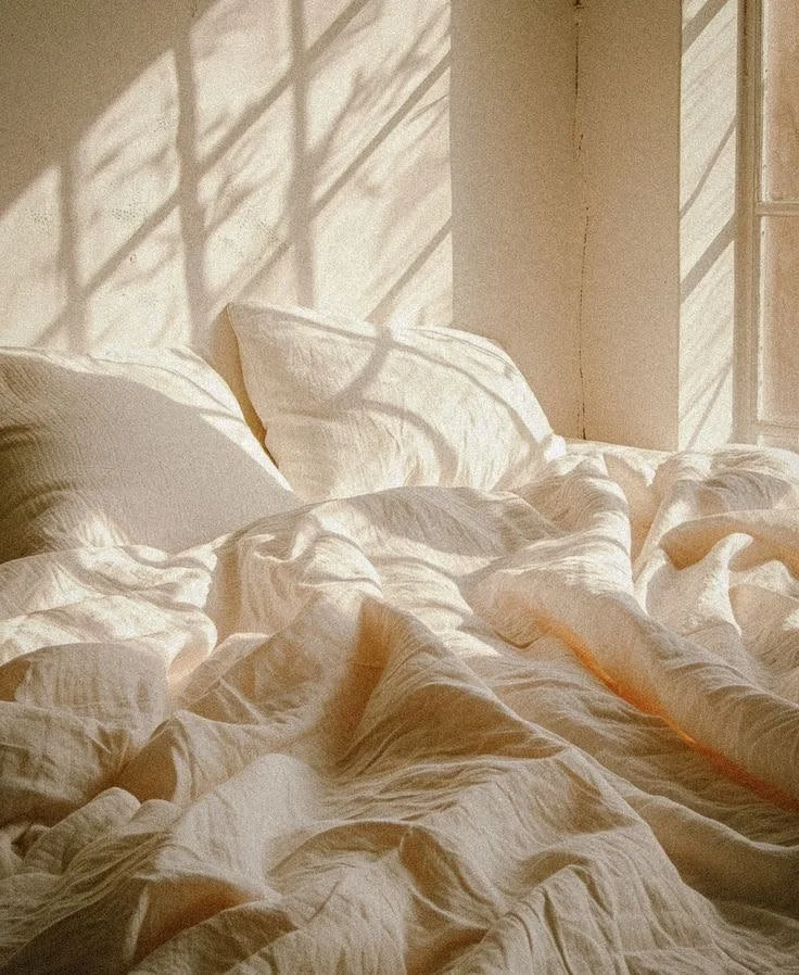 Sunlight casting shadows through window blinds onto an unmade bed with white sheets and pillows.
