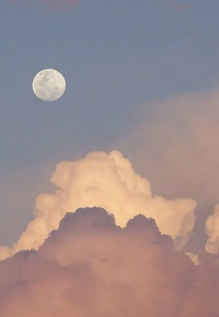 The moon in a clear sky above large, fluffy clouds.