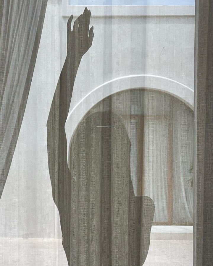 The shadow of a person with their arm raised, seen through a sheer curtain, with a window in the background.