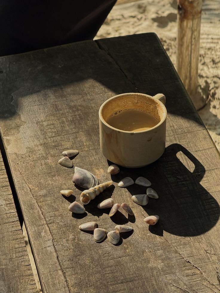 A beige mug filled with coffee on a weathered wooden surface, surrounded by an arrangement of small and large seashells, with a shadow cast from the mug.
