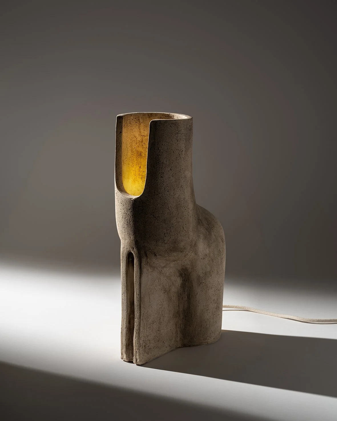 A tall, abstract ceramic lamp with a rough, textured surface. The lamp has a hollow, yellow illuminated interior visible through a cutout on the upper part. It is positioned on a plain surface with a shadow cast to the right.