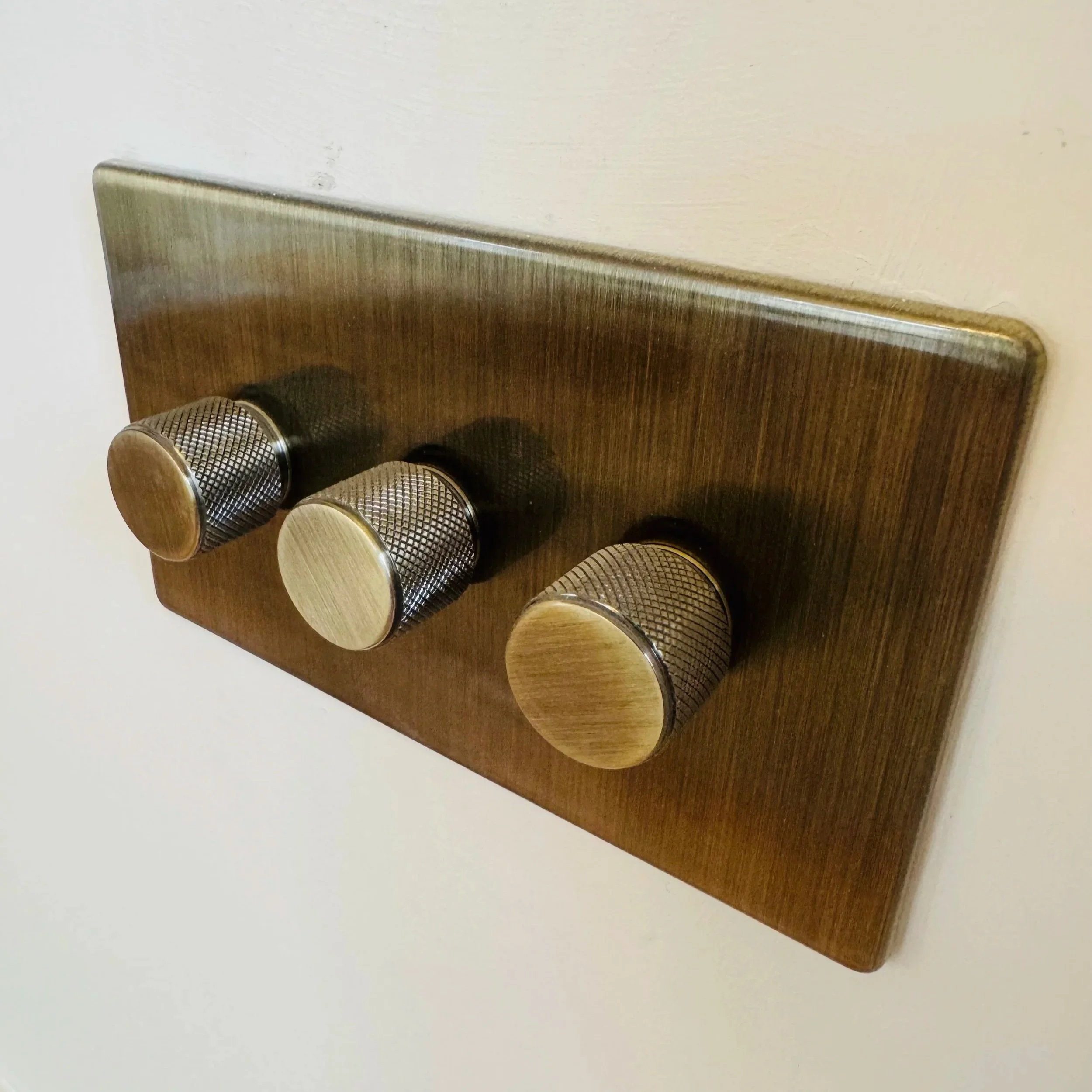 Close-up of a vintage light switch with three round, knurled metal knobs mounted on a brass plate on a white wall.