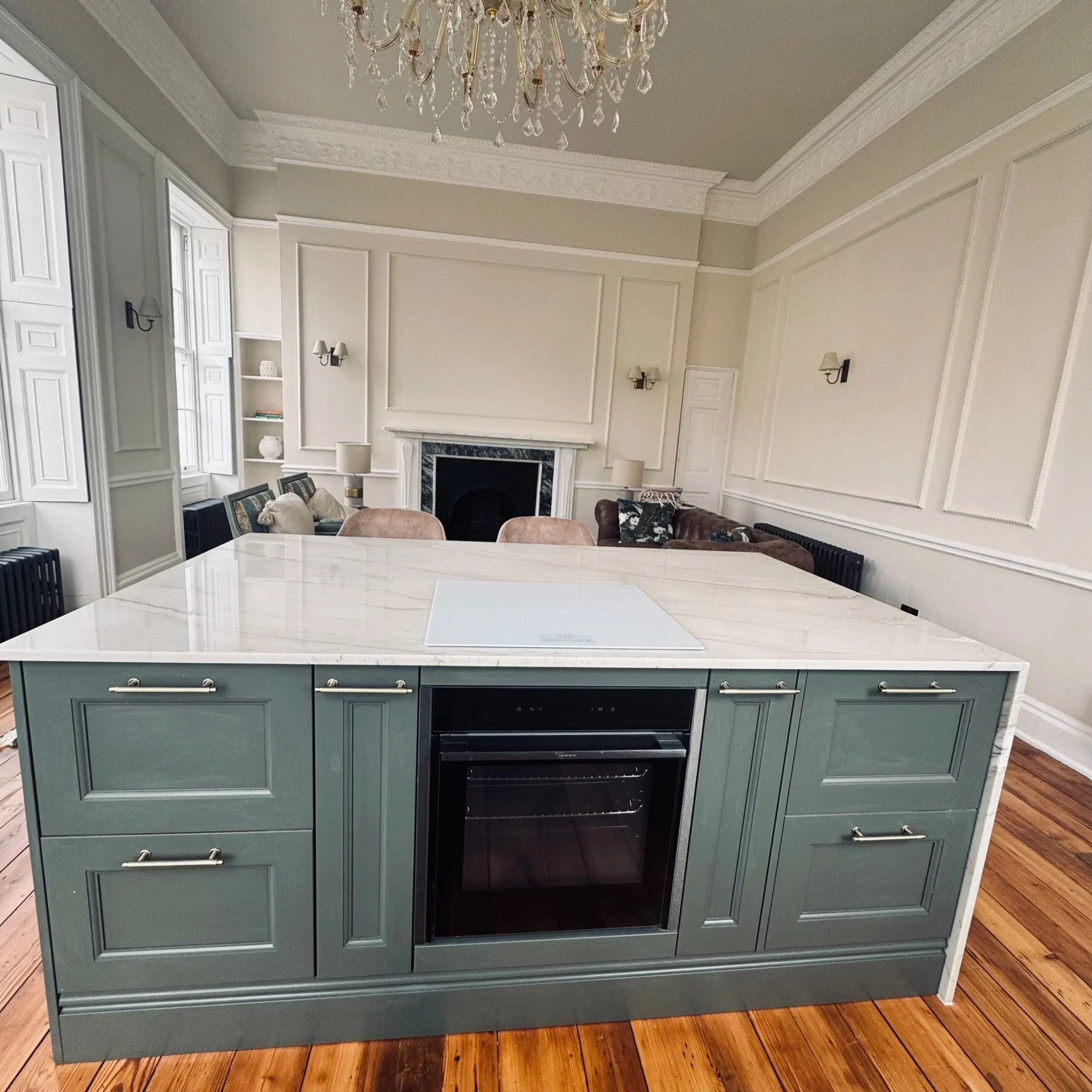 Modern kitchen island with a marble top, mint green cabinetry, and an oven, in a bright and spacious dining room with a chandelier and a fireplace.