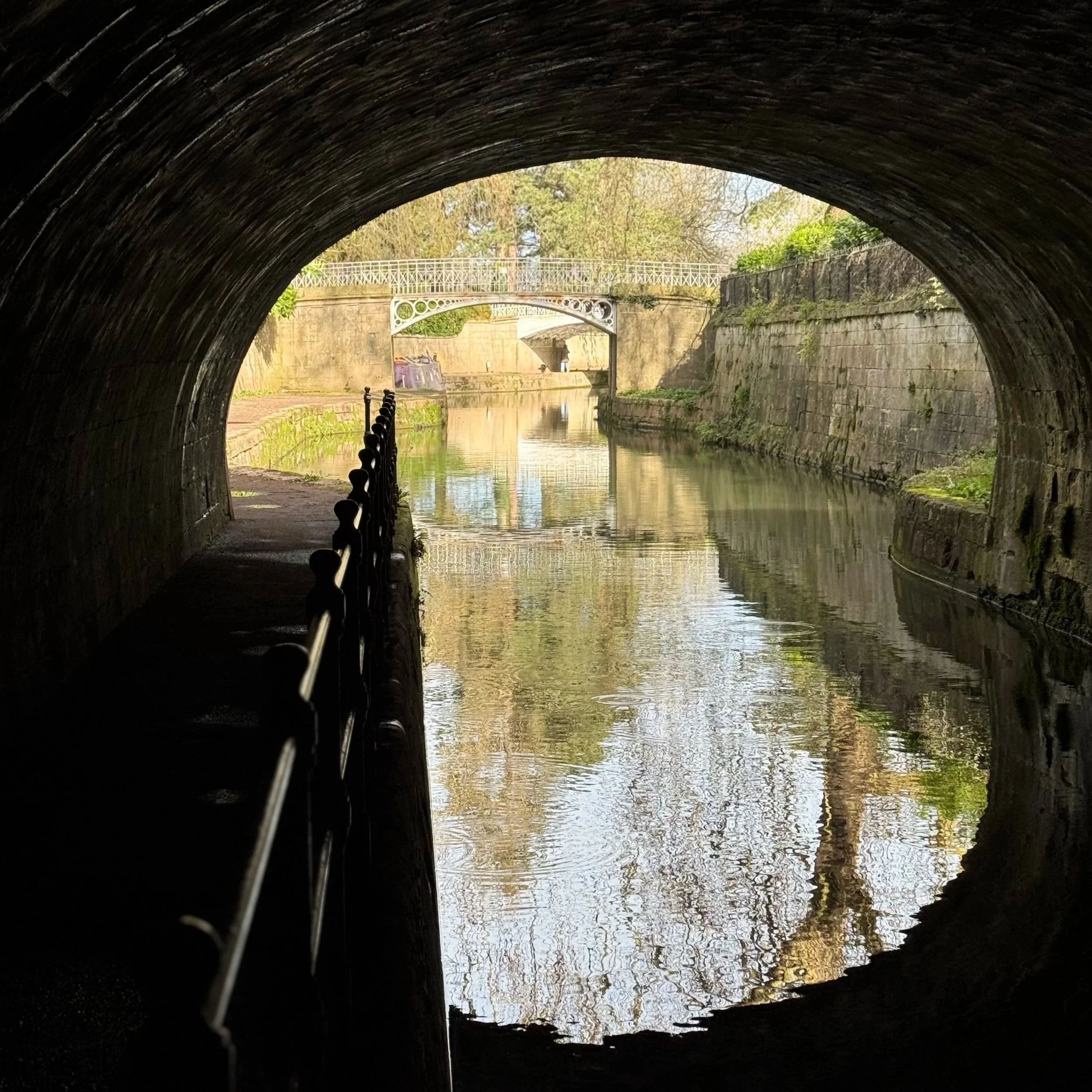 View from inside a tunnel looking out onto a waterway with a bridge in the distance, trees, and blue sky reflected in the water.