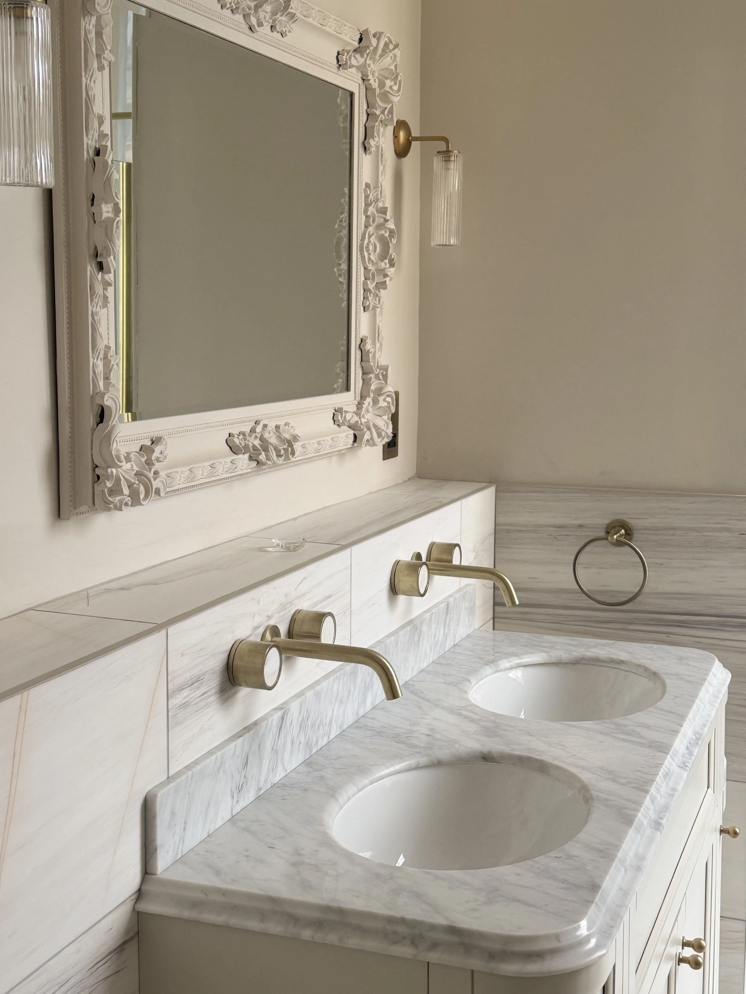 A bathroom vanity with a white marble countertop and two undermount sinks, featuring gold-colored fixtures. An ornate white-framed mirror hangs above, and a wall-mounted light fixture with a glass shade is beside it. There is also a towel ring to the