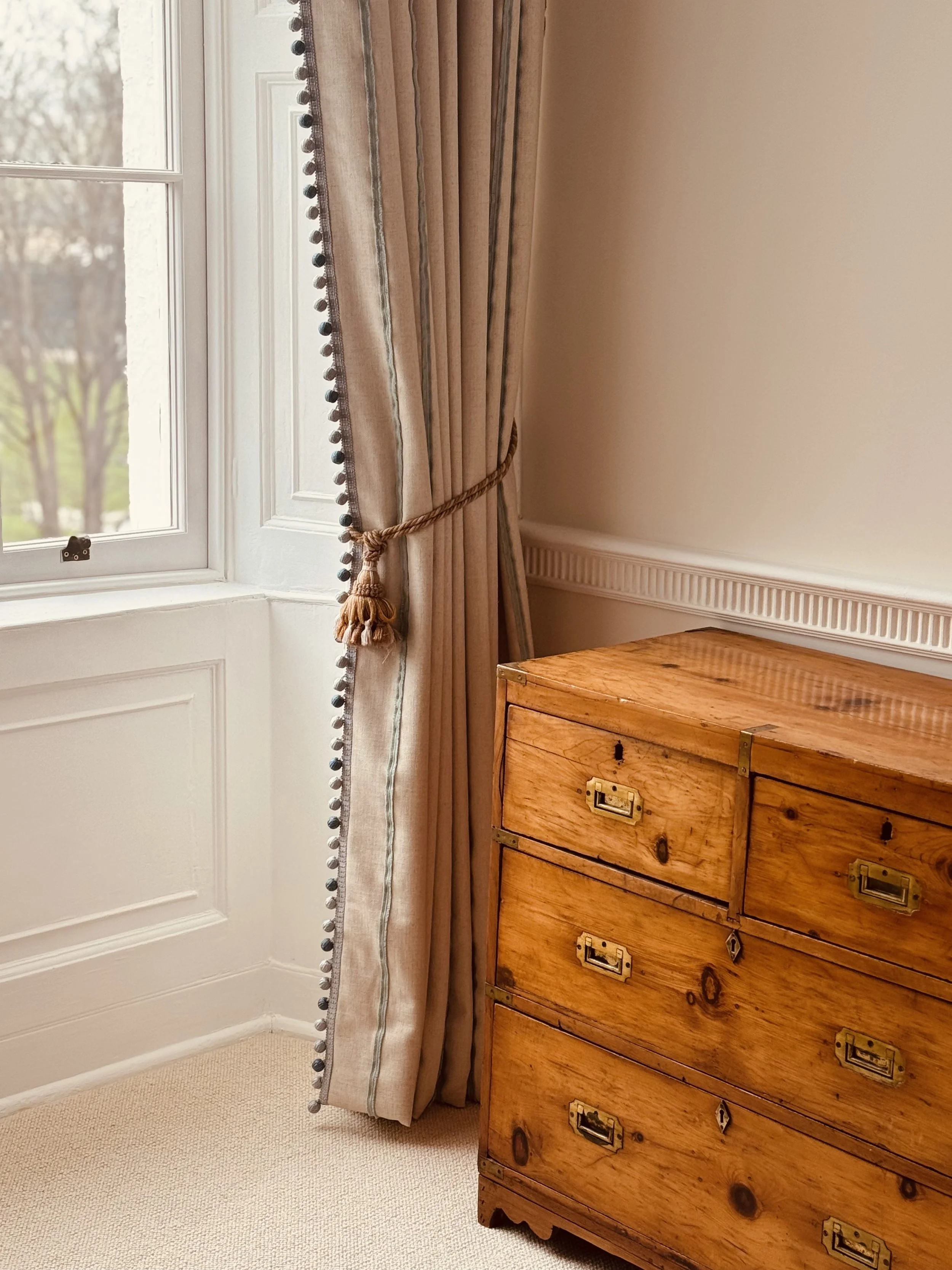A corner of a room with beige walls, a window with beige curtains tied back with a rope, and a wooden dresser with multiple drawers.