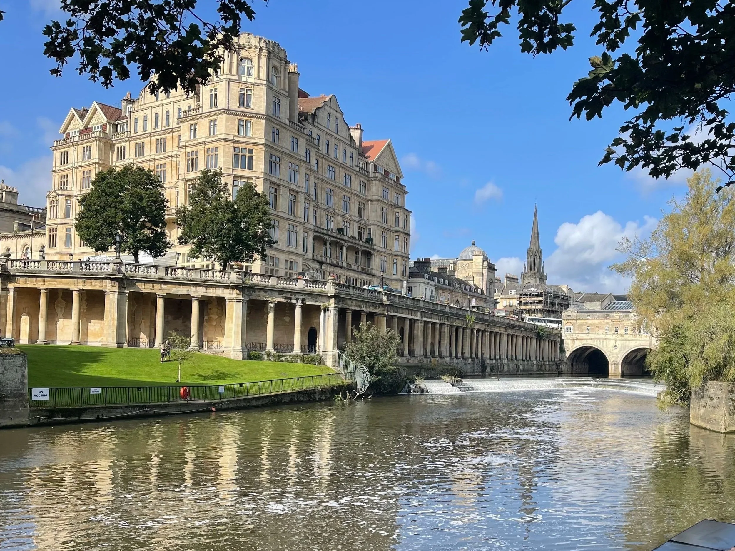 View of a historic Bath stone building overlooking Pultney Bridge weir under a blue sky with clouds, surrounded by trees with some green foliage.