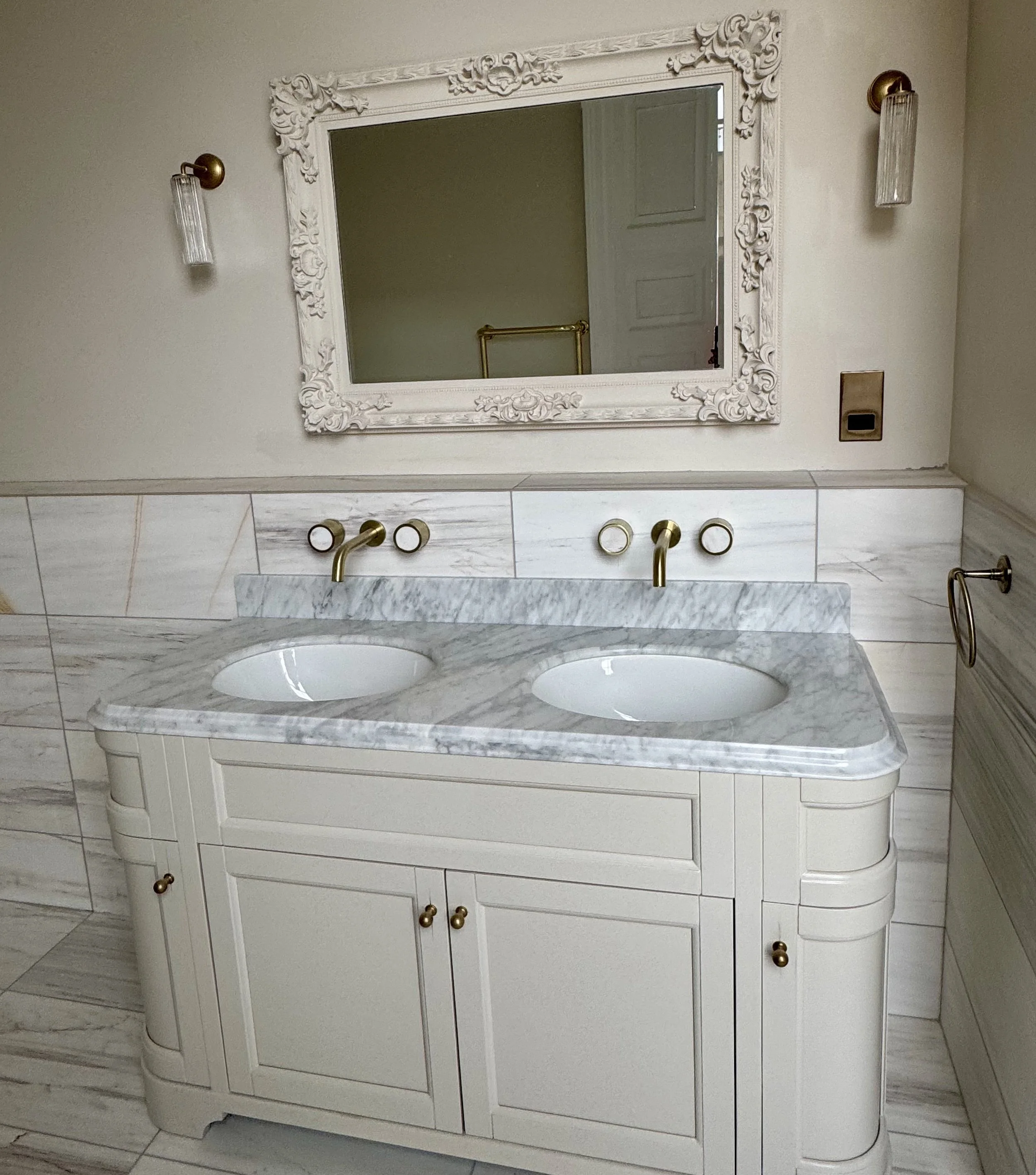 Bathroom vanity with a marble countertop, double sinks, gold fixtures, a large ornate mirror, and wall-mounted lights.