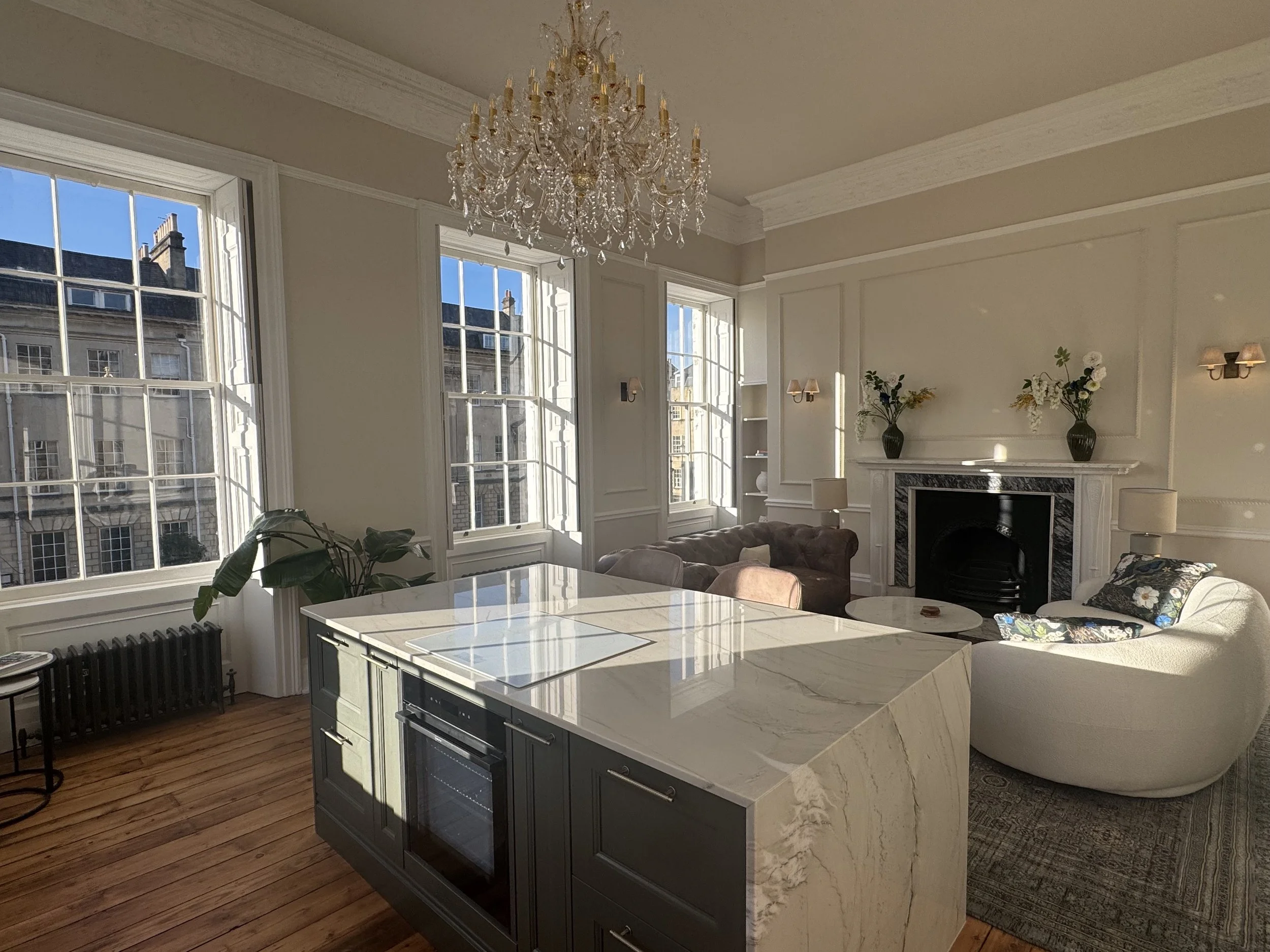 Living room with large windows, chandelier, marble fireplace with vases and flowers, several antique armchairs, and a marble kitchen island.