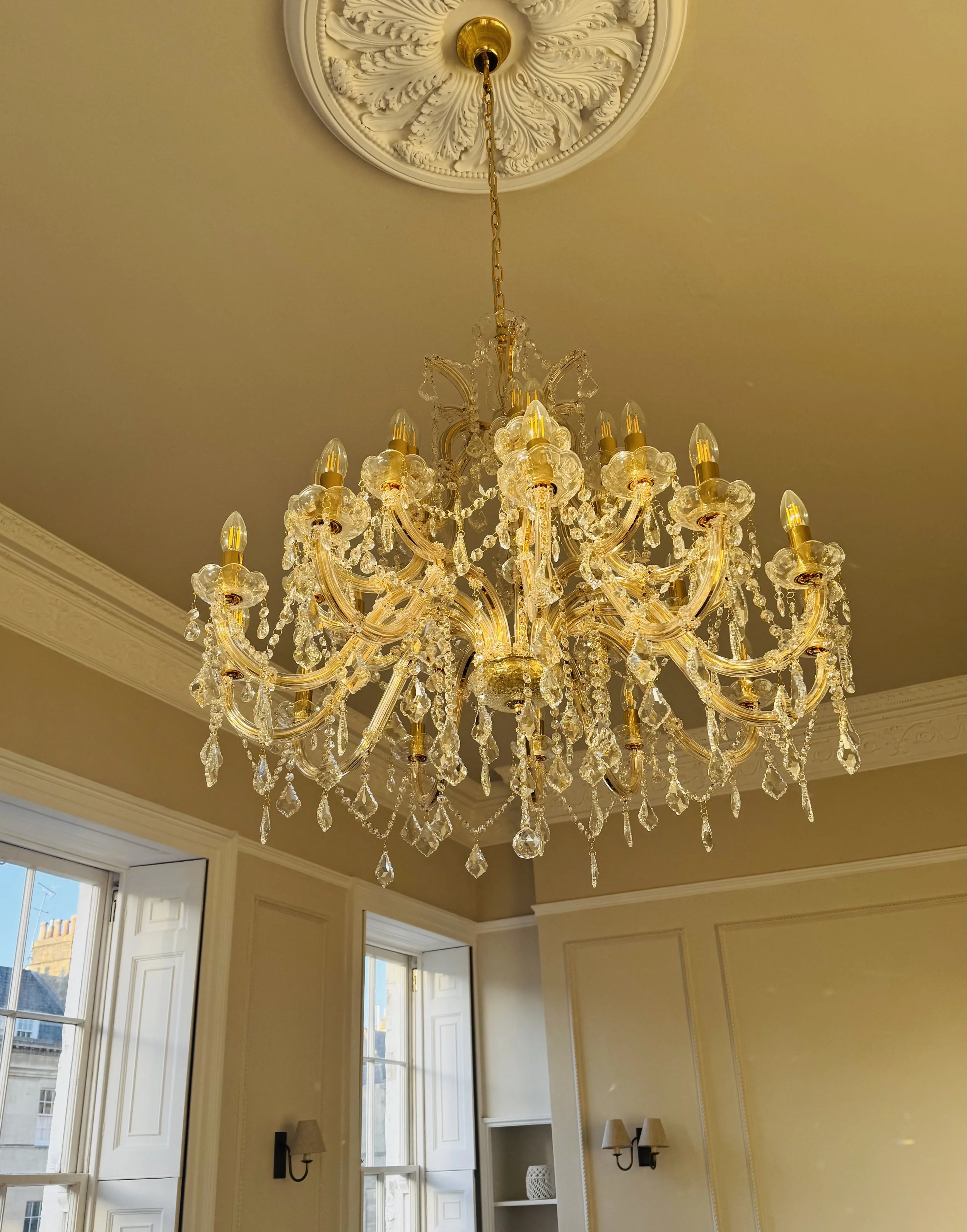 An ornate gold and crystal chandelier hanging from a decorative ceiling medallion in a well-lit room with cream-colored walls and large windows.