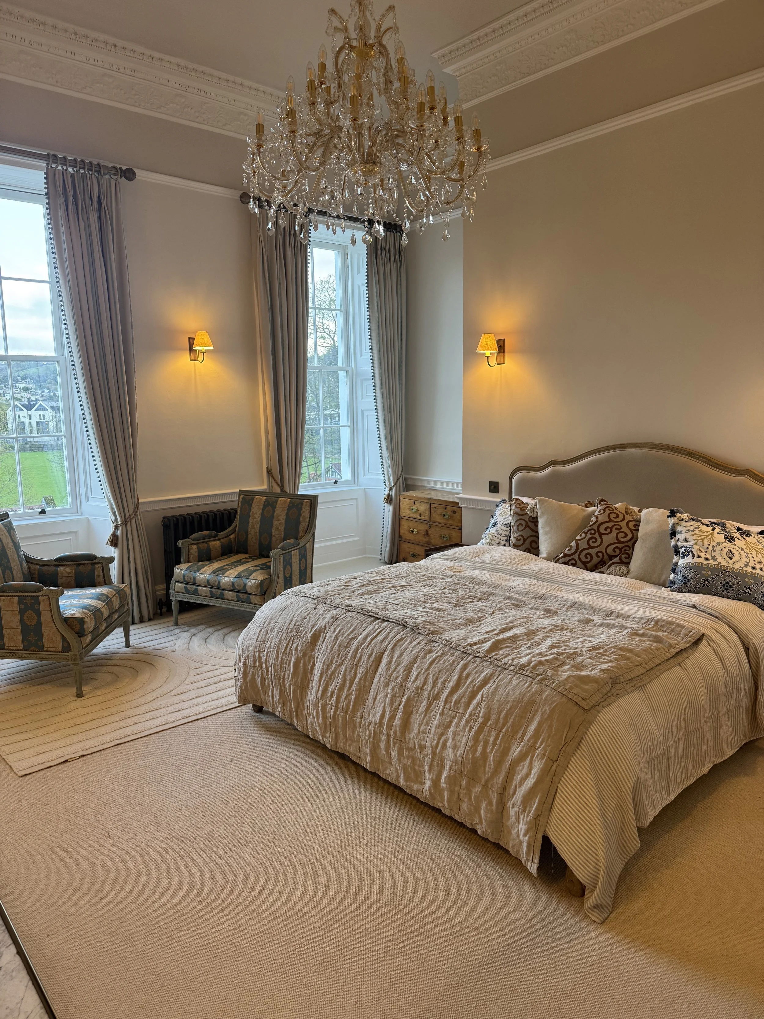 Elegant bedroom with large bed, chandelier, two antique armchairs, windows with bespoke curtains, wall sconces, and an antique dresser.