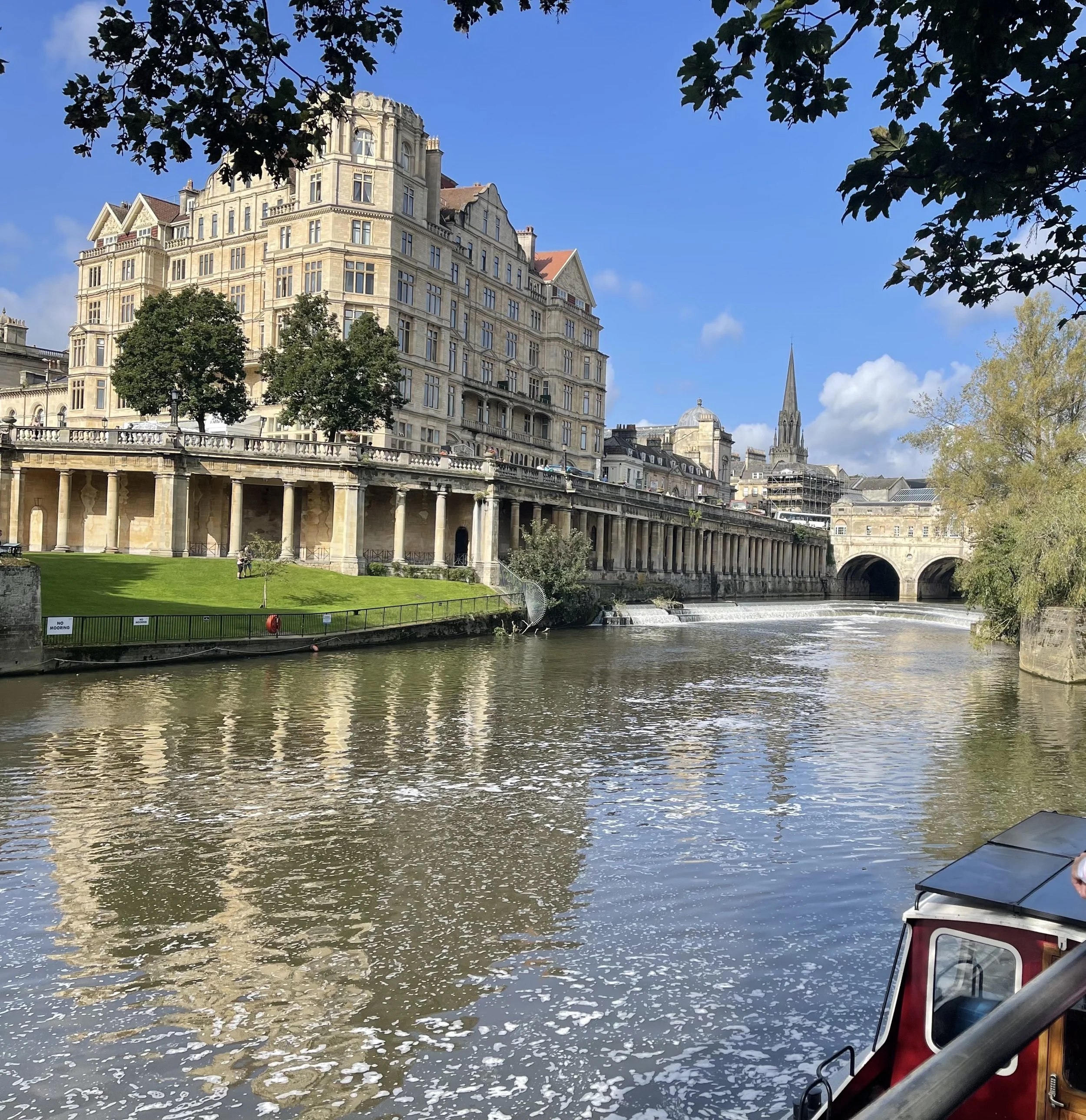 View of a river with a building and a church steeple in the background, trees along the riverbank, and a boat partially visible in the bottom right corner.