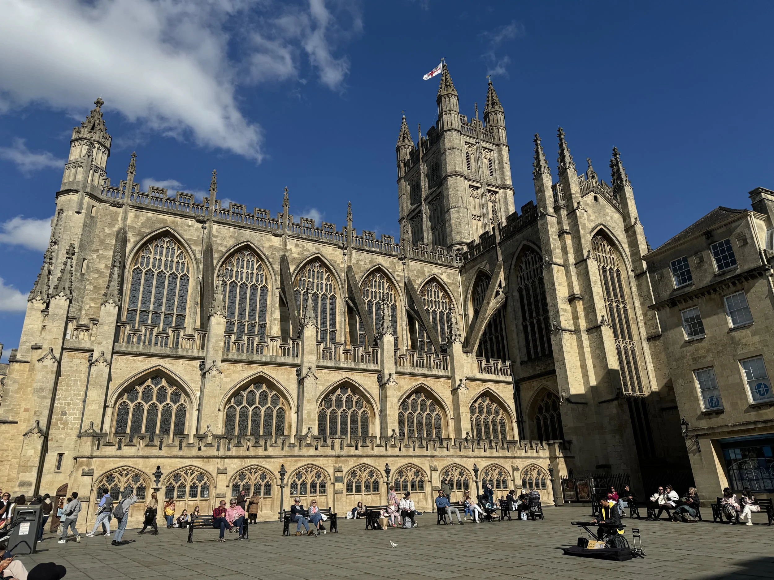 People sitting and walking outside of a large, historic Gothic Abbey with tall windows, flying a flag on a tower, against a blue sky with clouds.
