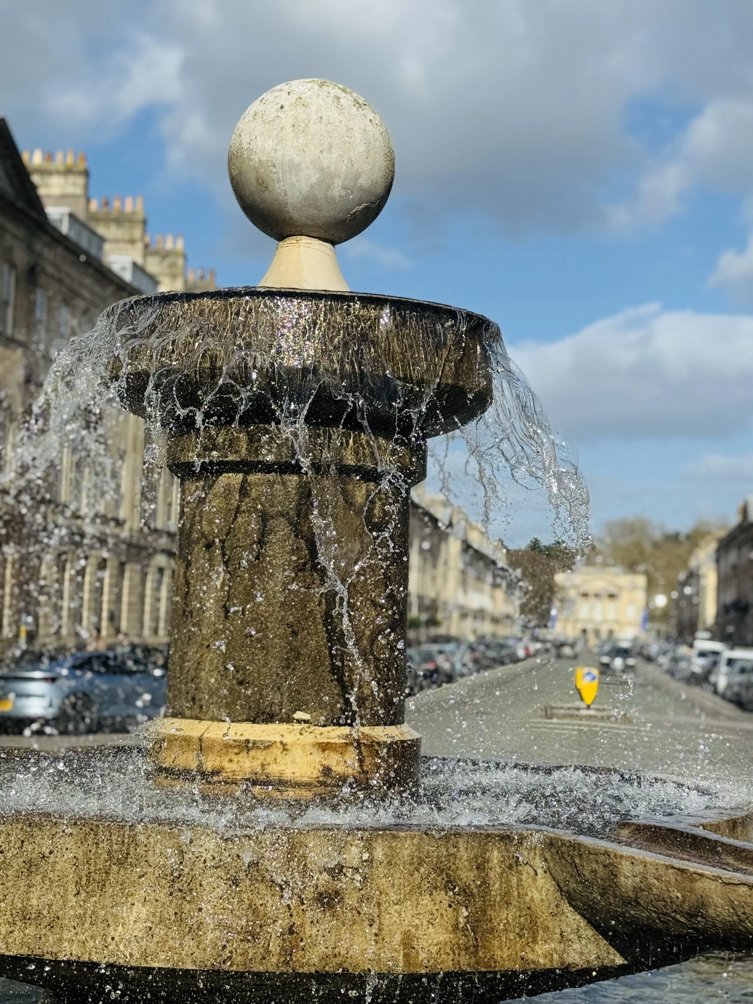 Close-up of a stone fountain with water flowing from its edges, set in an urban area with buildings and parked cars in the background, under a partly cloudy sky.