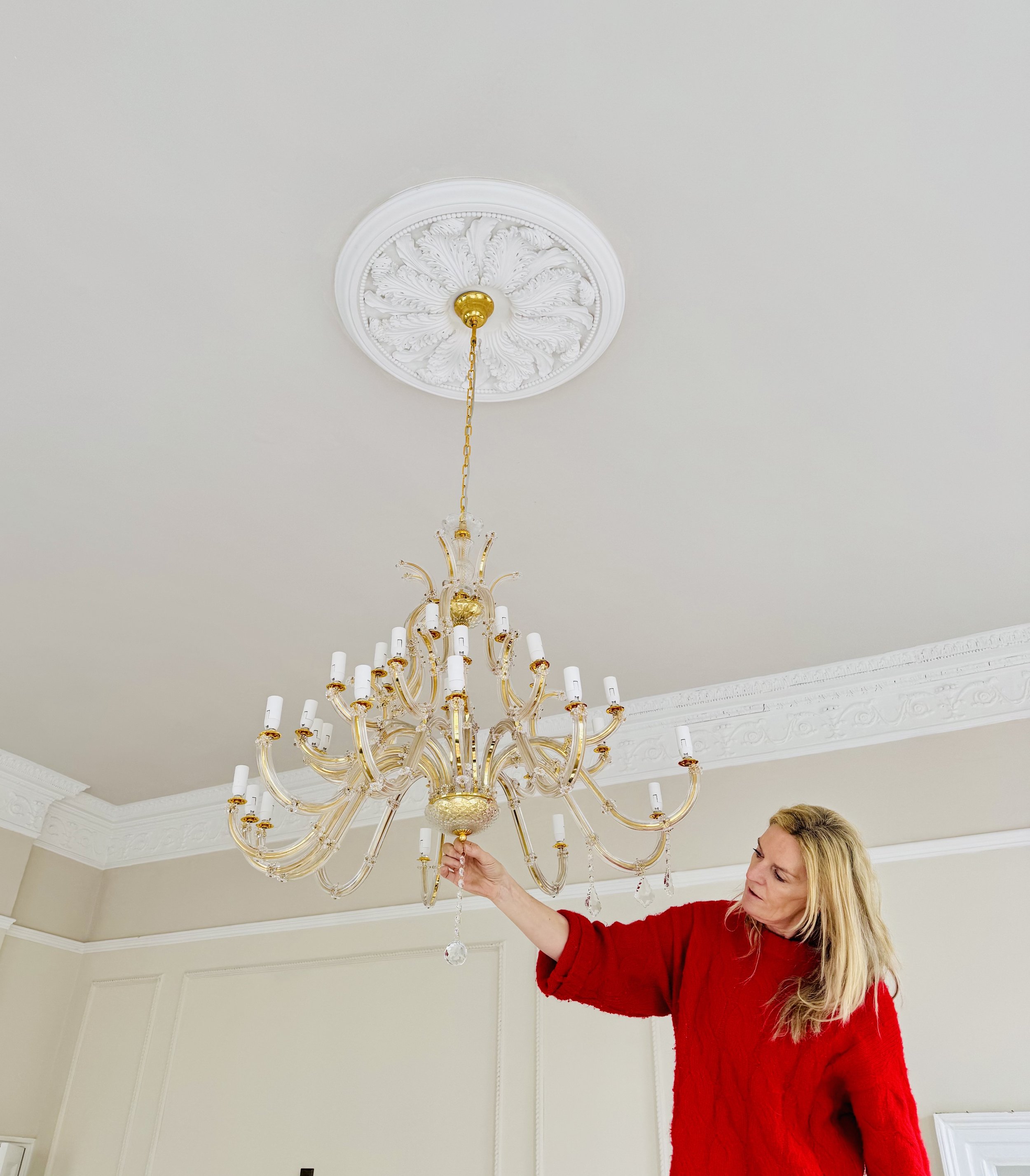Queen Charlotte of Bath in red sweater adjusting a large decorative gold chandelier in a room with ornate crown molding.