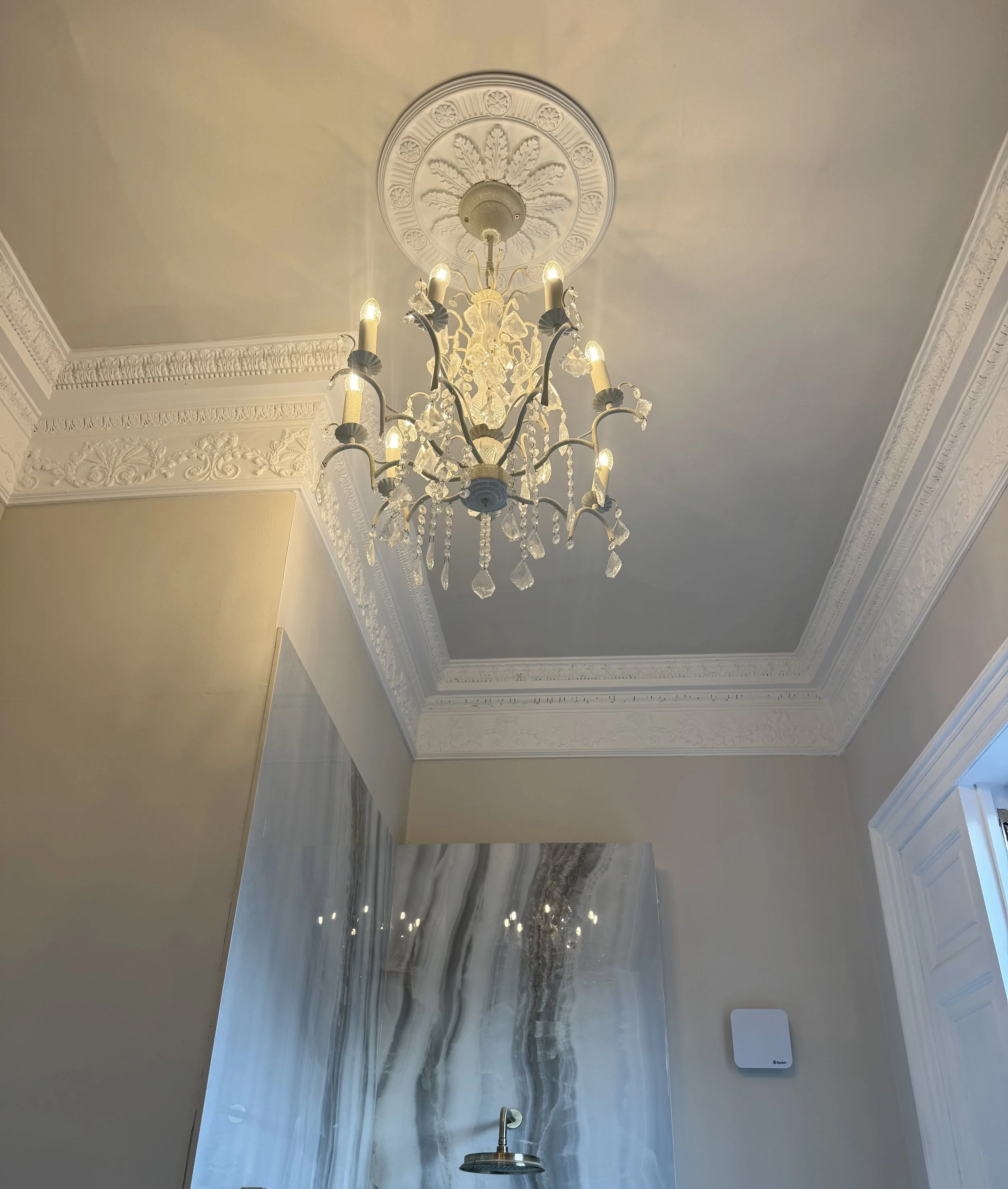 A ceiling chandelier with multiple candle-shaped lights hanging from a decorative ceiling medallion, surrounded by ornate crown molding in a room with beige walls and a marble feature wall.