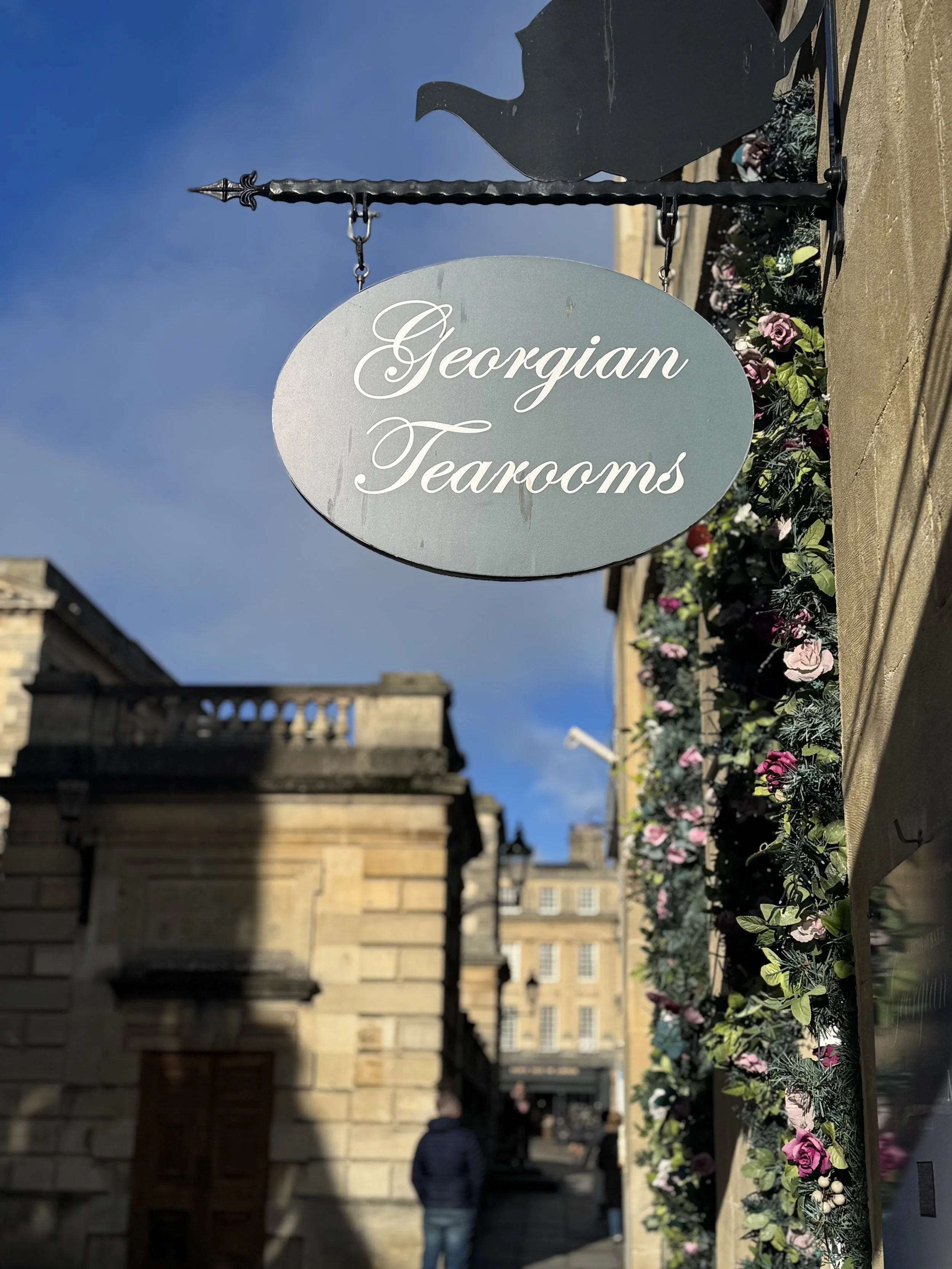 Sign for Georgian Tearooms hanging outside with a city street and historic buildings in the background, decorated with flowers.