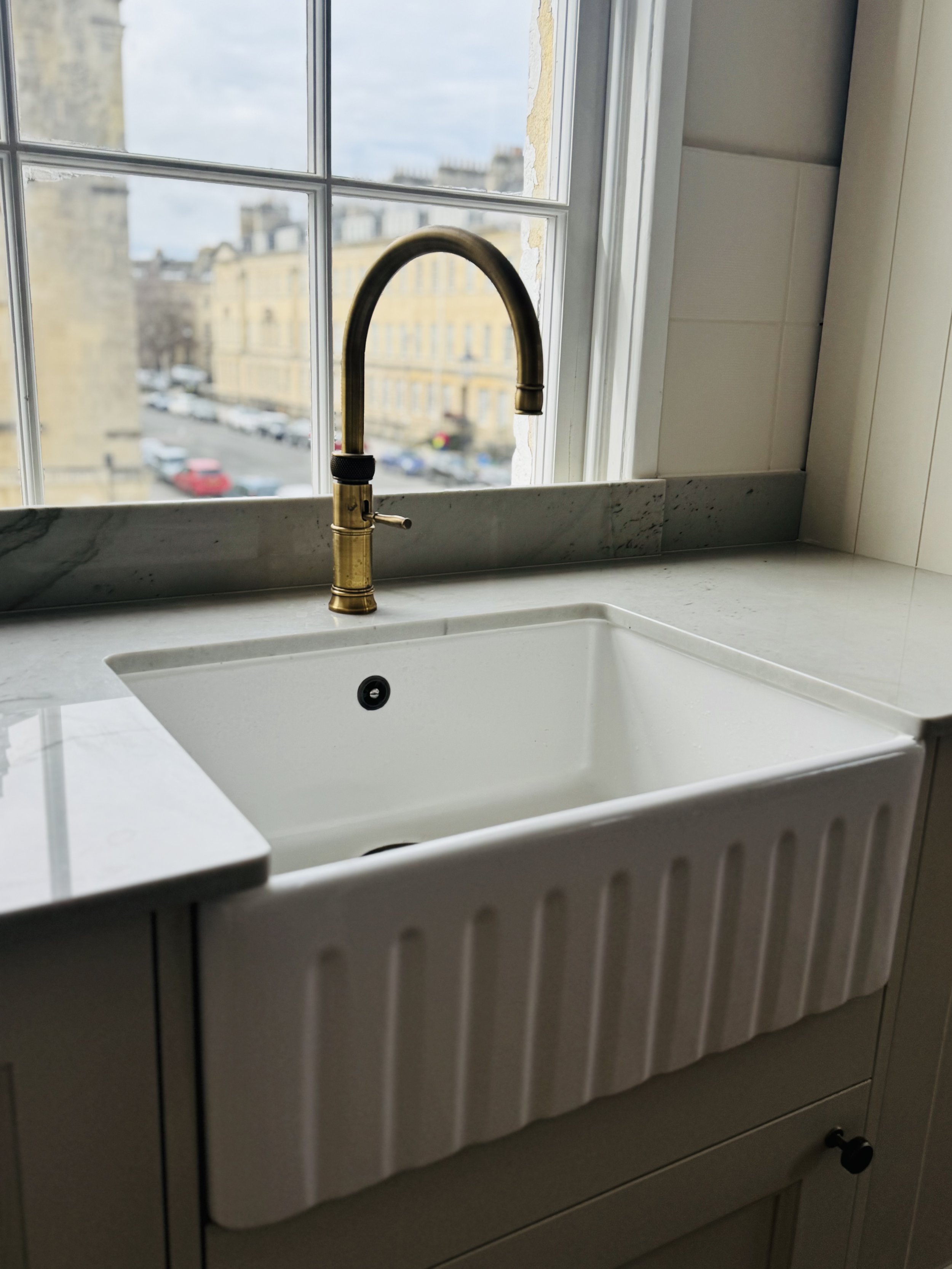A white kitchen sink with a brass faucet, set against a window overlooking a city street with parked cars and beige buildings.