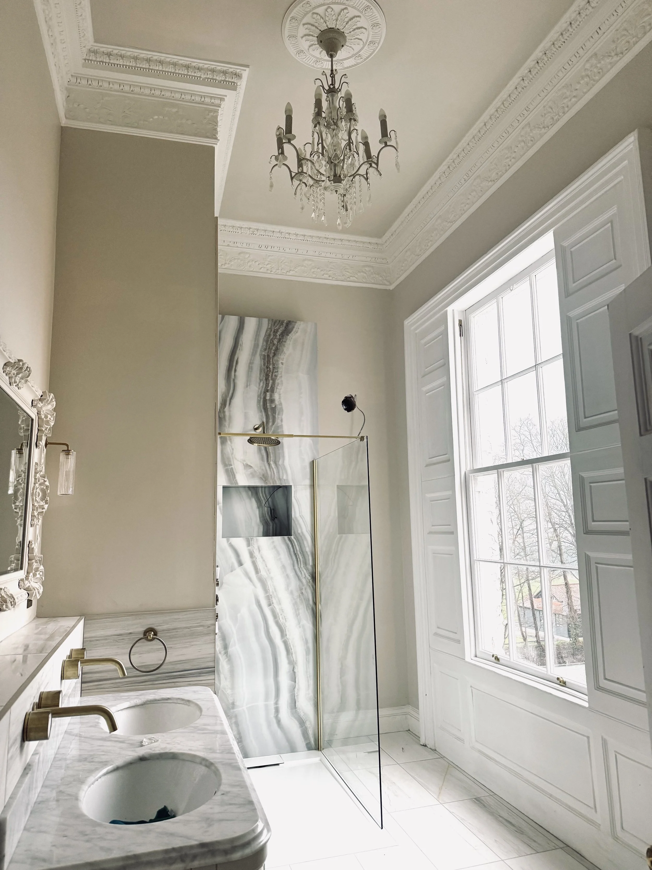 Elegant bathroom with high ceilings, ornate crown molding, a crystal chandelier, a large window with white trim, a marble vanity with two sinks, a marble shower wall with gold accents, and a glass shower door.