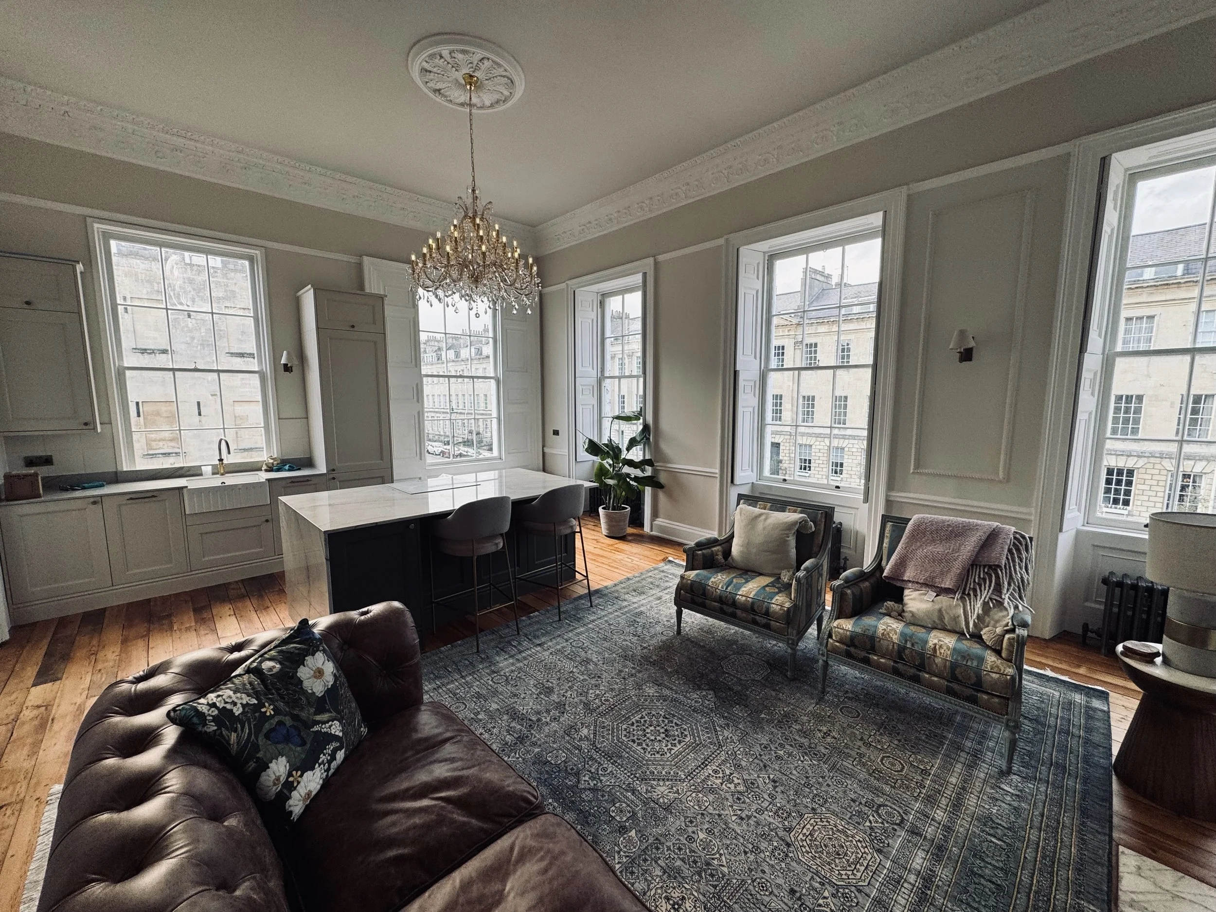 Elegant living room with large windows, a chandelier, a leather sofa with floral pillow, two patterned armchairs with blankets, a rug, and a kitchen island with bar stools.