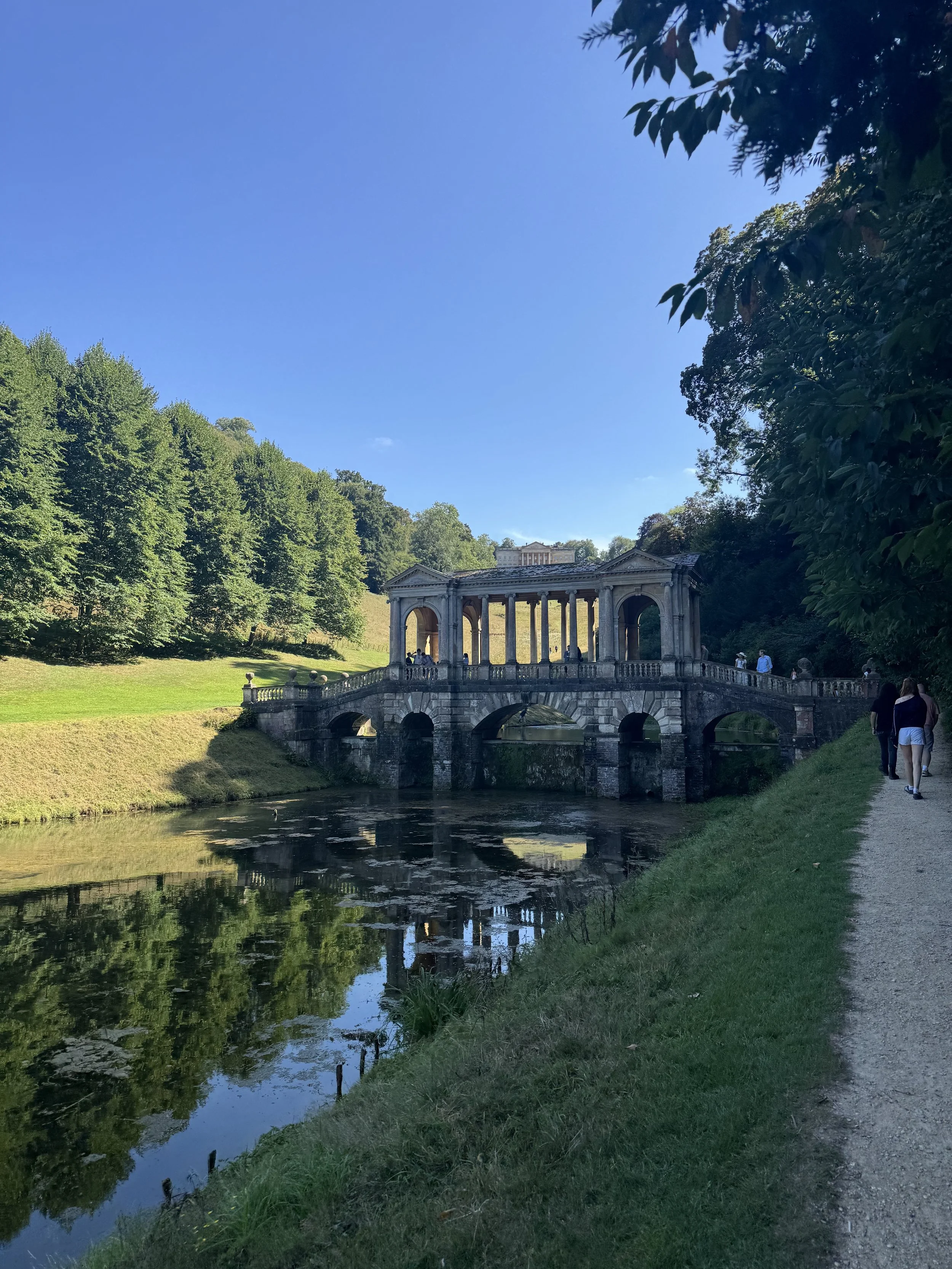 Palladian bridge with classical columns over a small river in a park, with trees, grass, and people walking on a path under a blue sky.