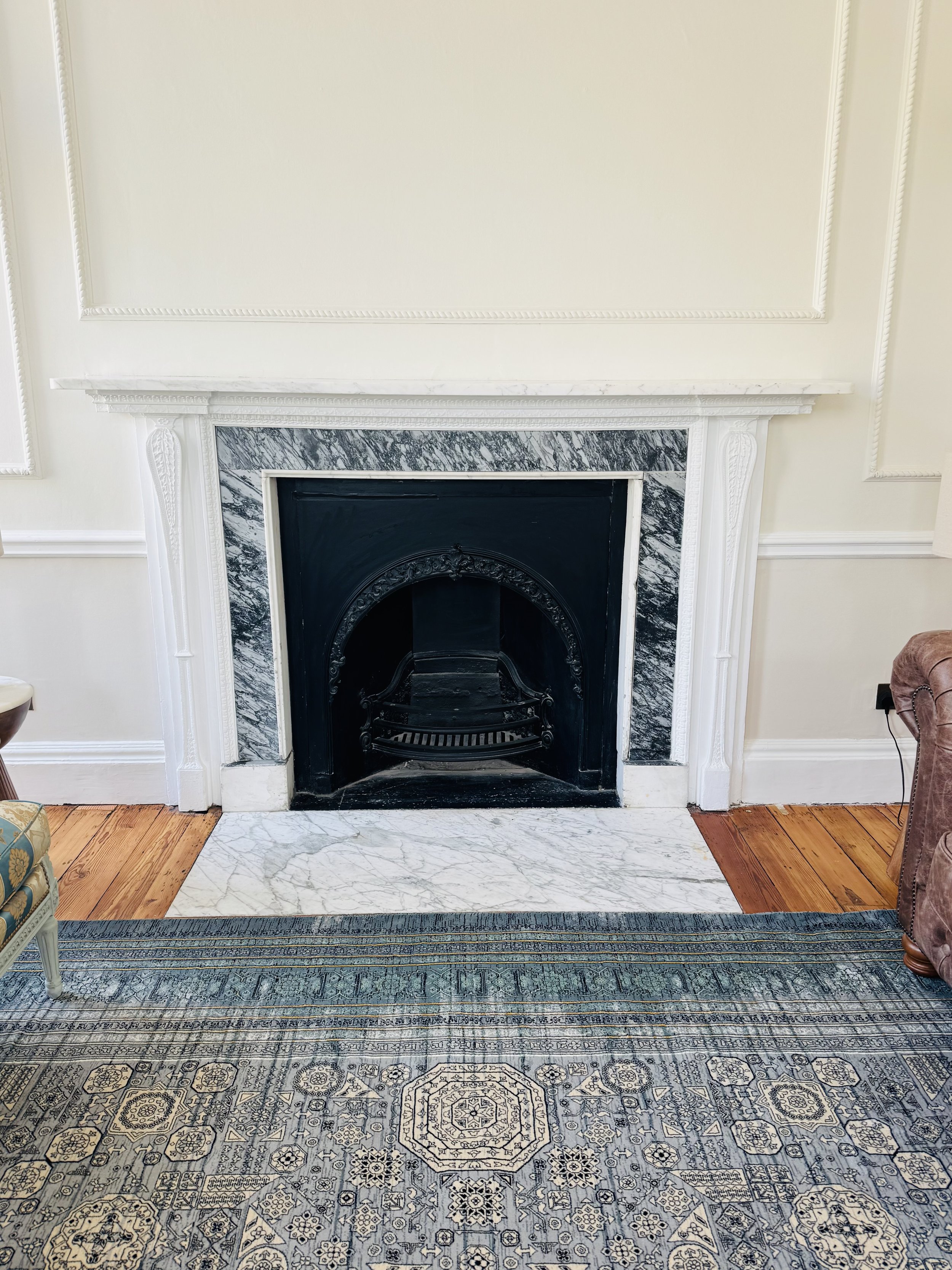 Living room with white fireplace, black interior, marble and wood flooring, patterned blue and beige area rug, partially visible armchairs.