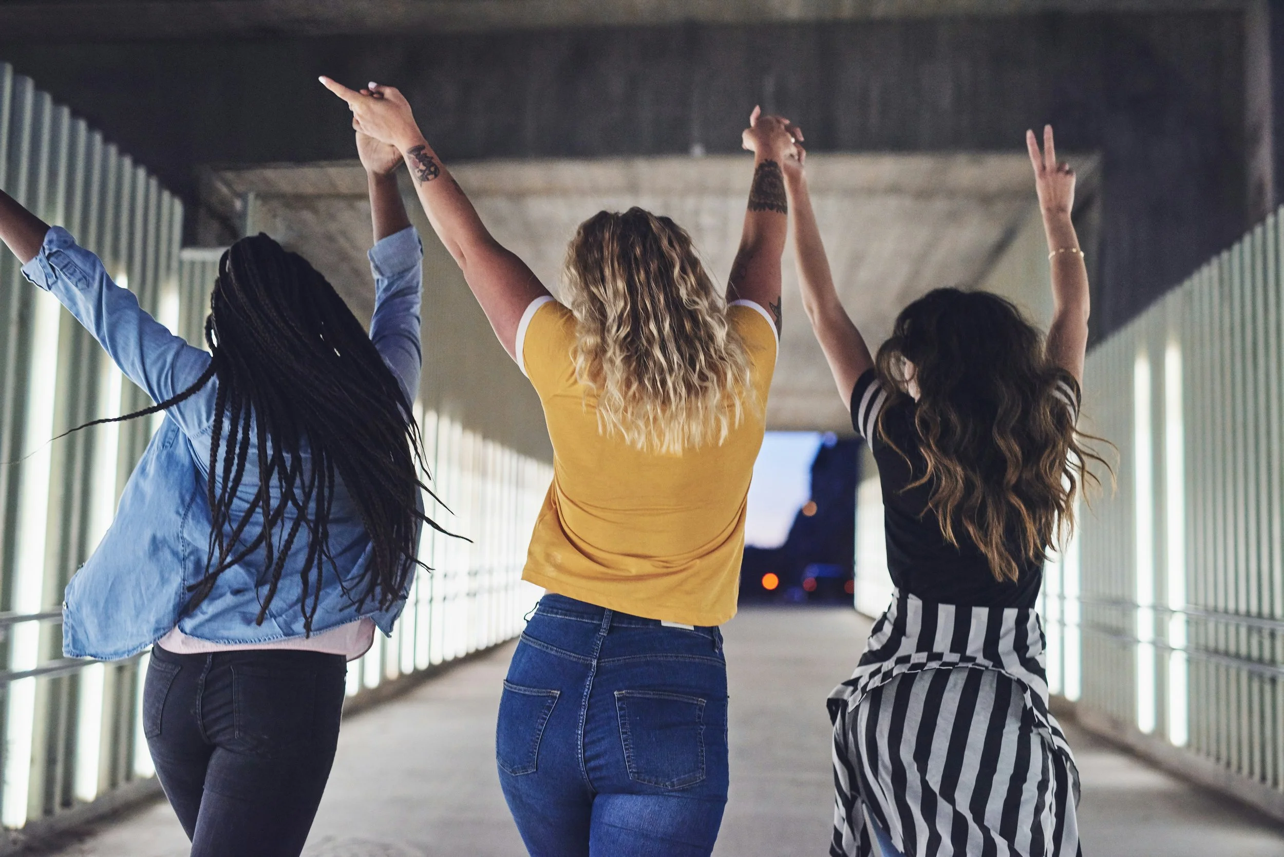 Three women standing in an underpass, raising their arms and making peace signs or pointing while facing away from the camera.