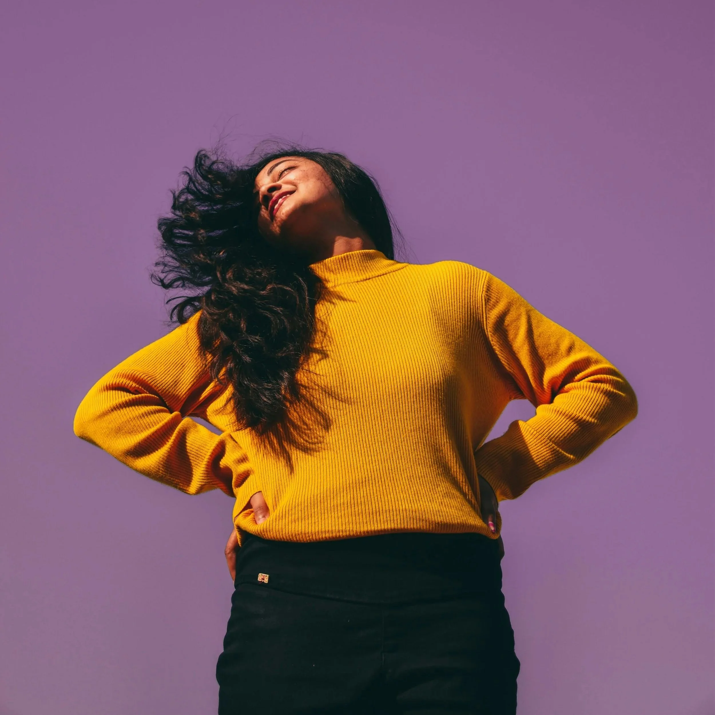 A person wearing a yellow sweater and black pants standing against a purple background, with long curly hair and a big smile.