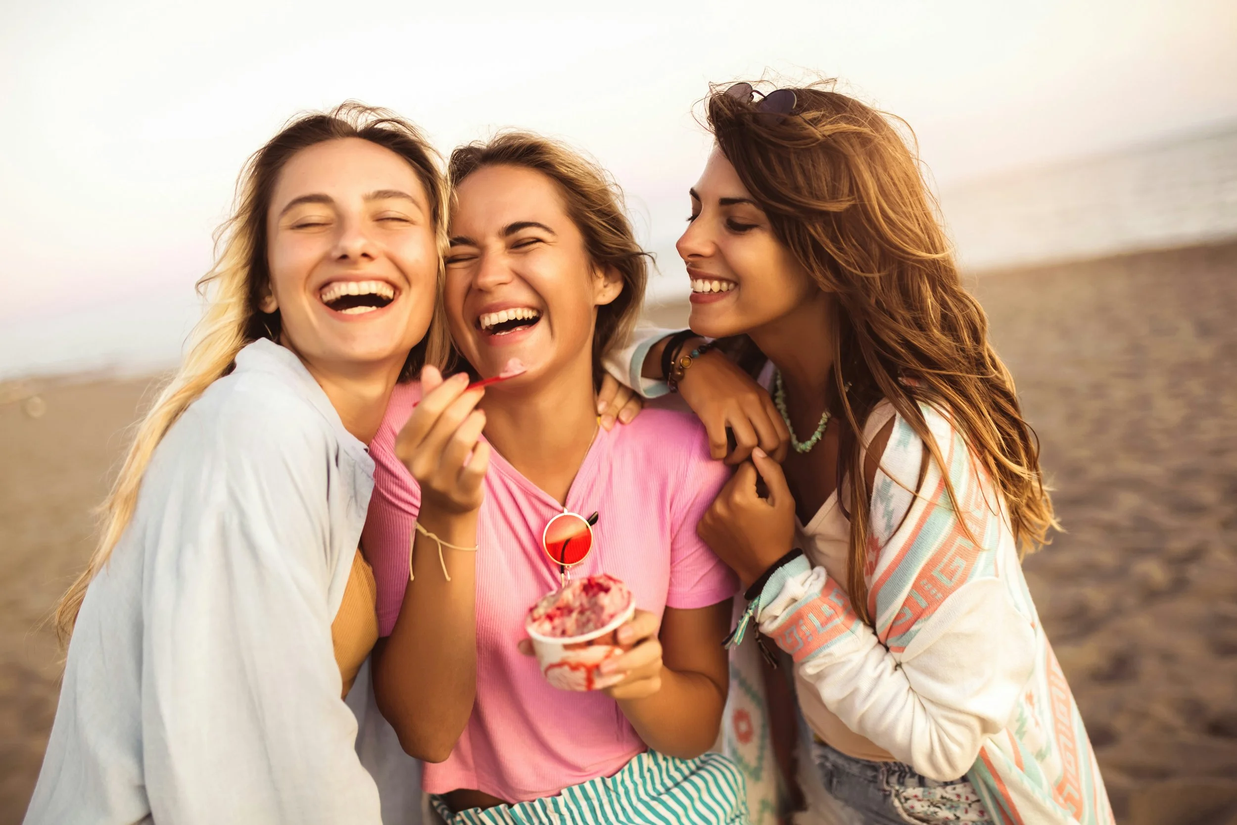Three young women laughing and hugging each other at the beach, one is holding a cup of ice cream.
