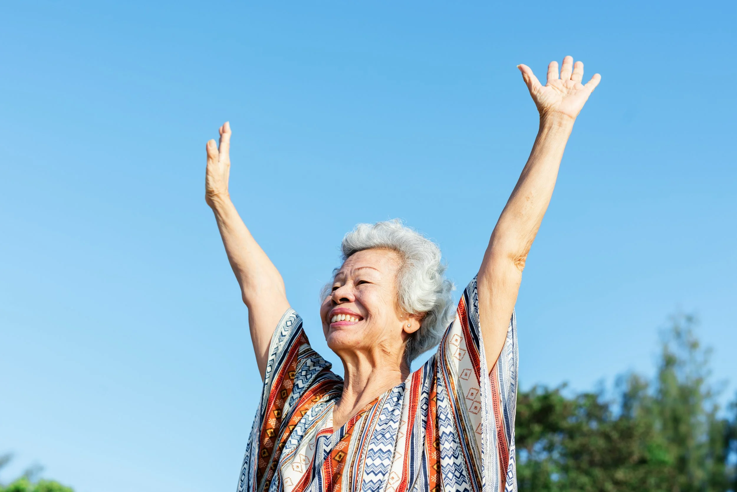An elderly woman with gray hair smiling and raising her arms in the air outdoors against a clear blue sky.