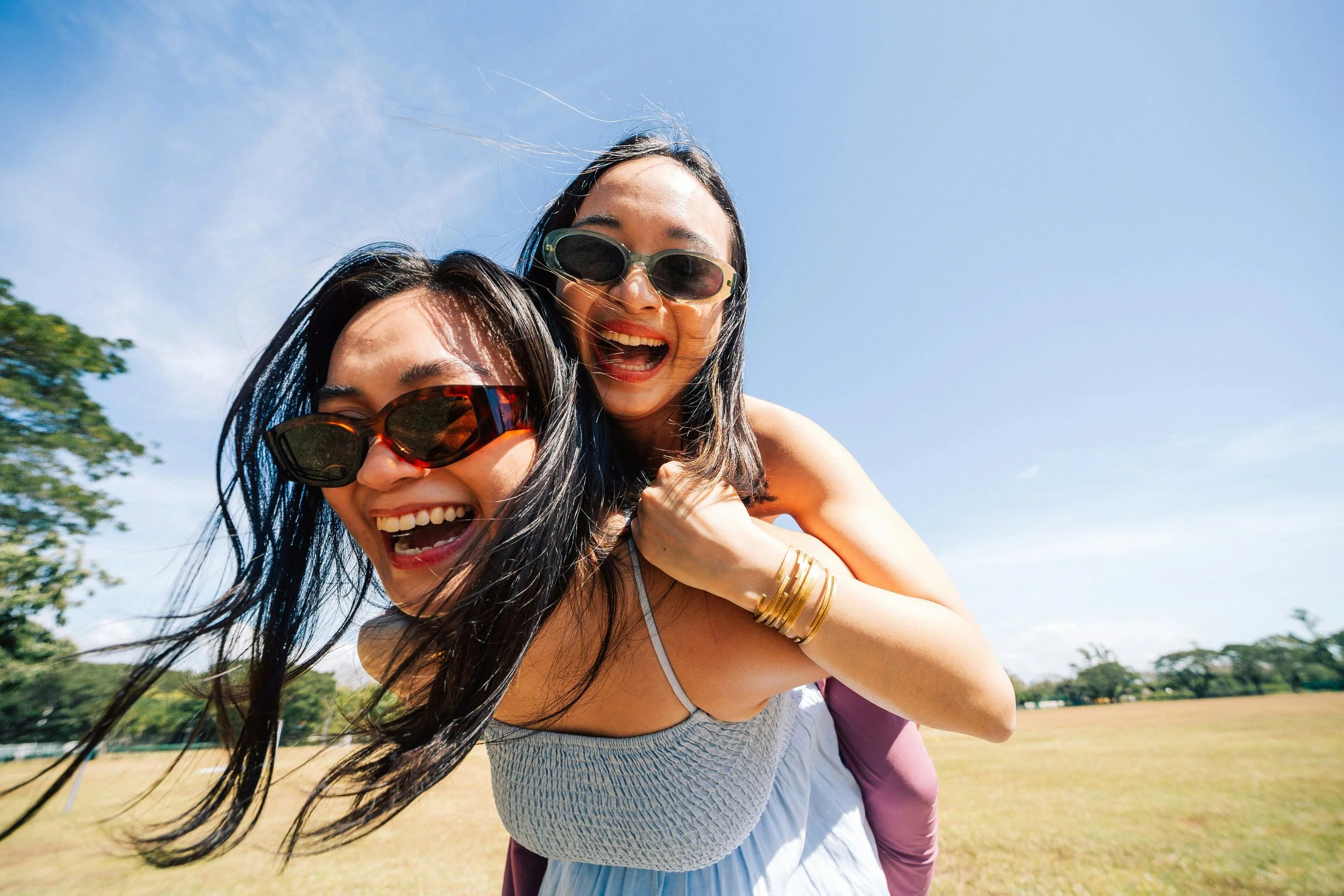 Two women with dark hair and sunglasses smiling outdoors on a sunny day, with trees and a grassy field in the background.