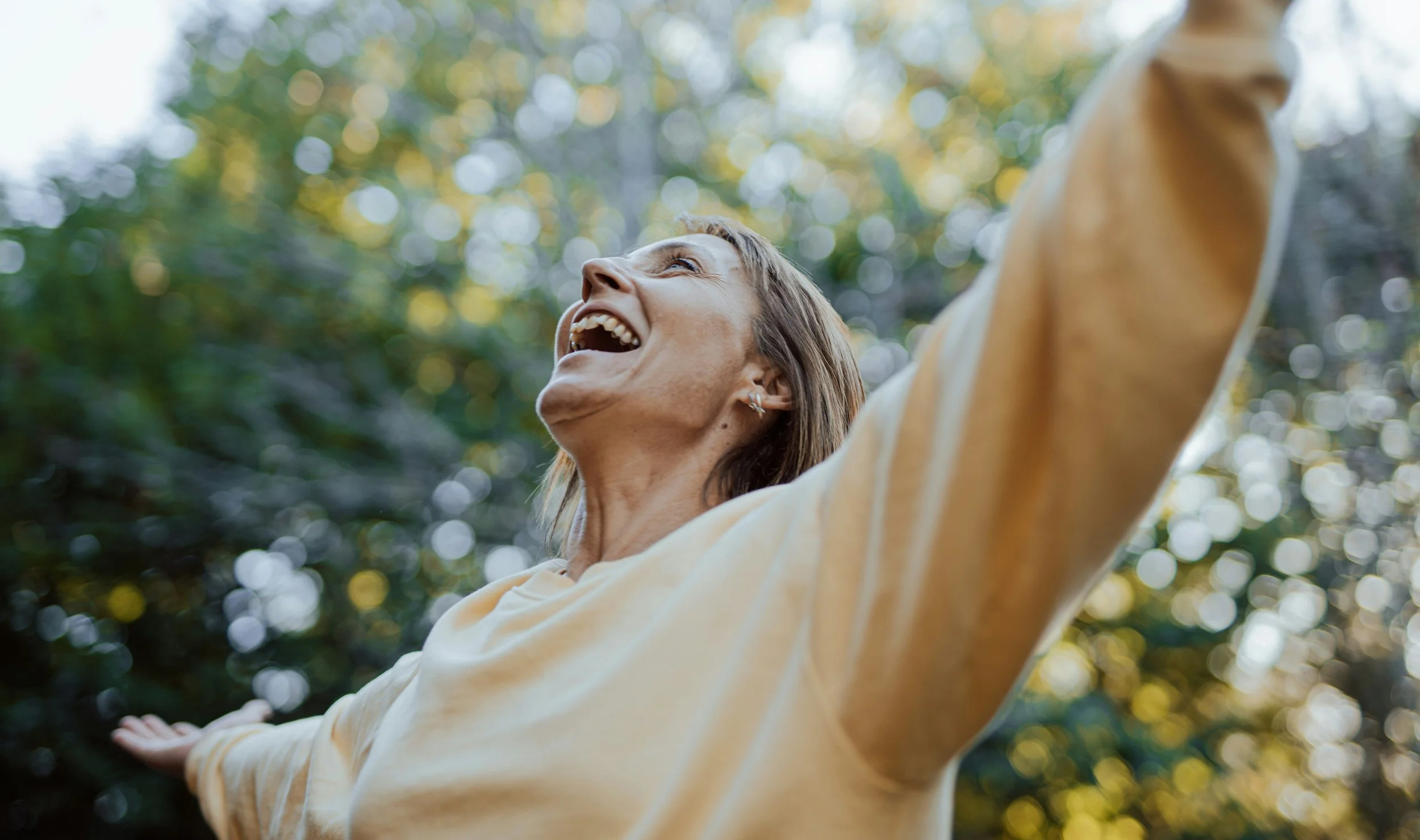 A woman outdoors, smiling and raising her arms, with sunlight filtering through trees in the background.