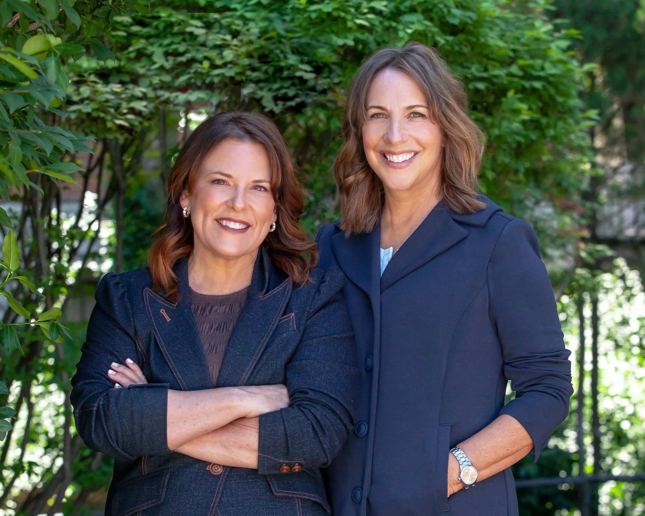 Two women standing outdoors near green foliage, smiling at the camera. One woman has brown hair and is wearing a denim jacket, the other has light brown hair and is dressed in a navy blazer.