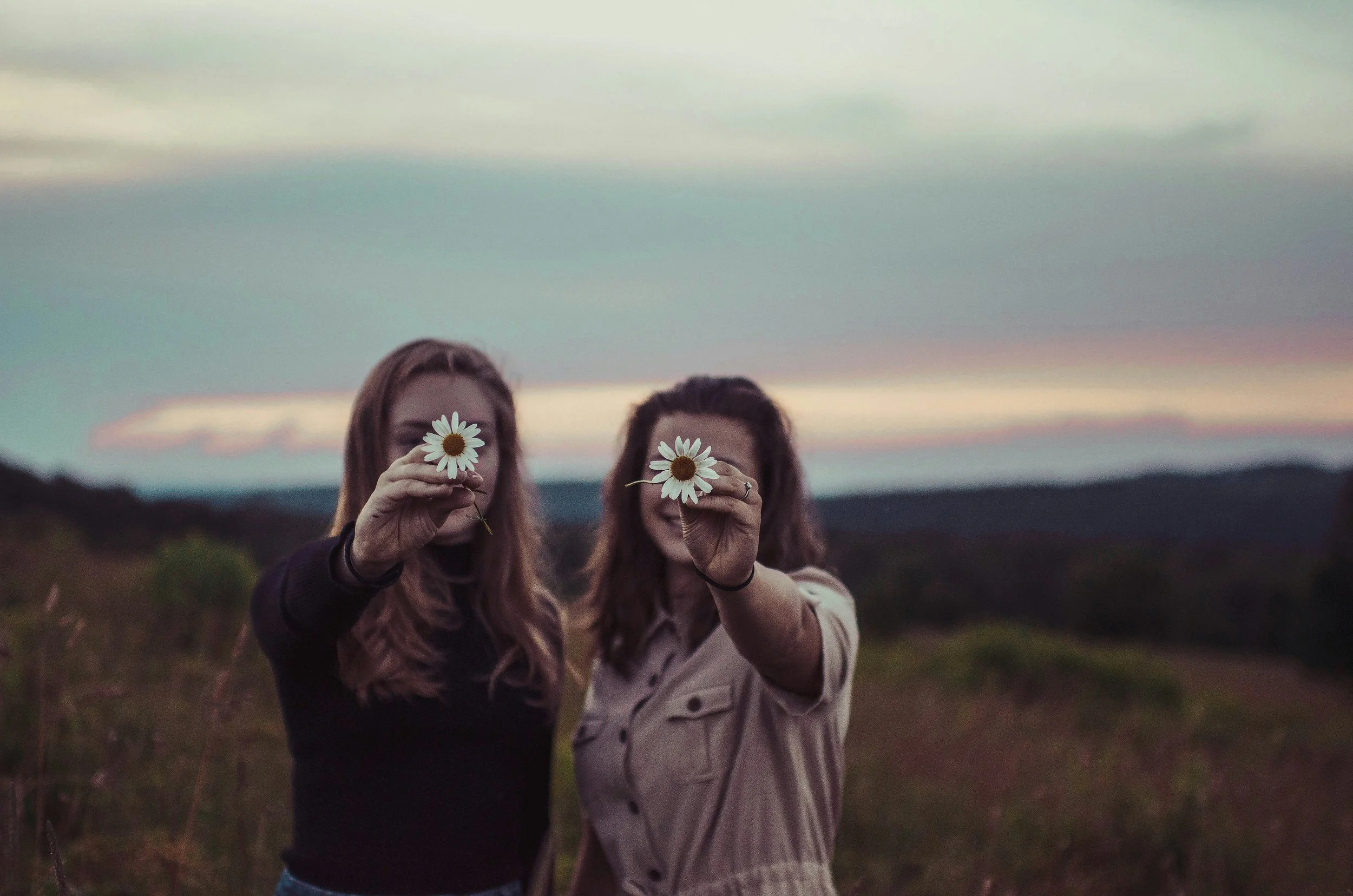 Two women holding daisies in front of their faces with a background of an open sky and distant landscape during sunset or sunrise.