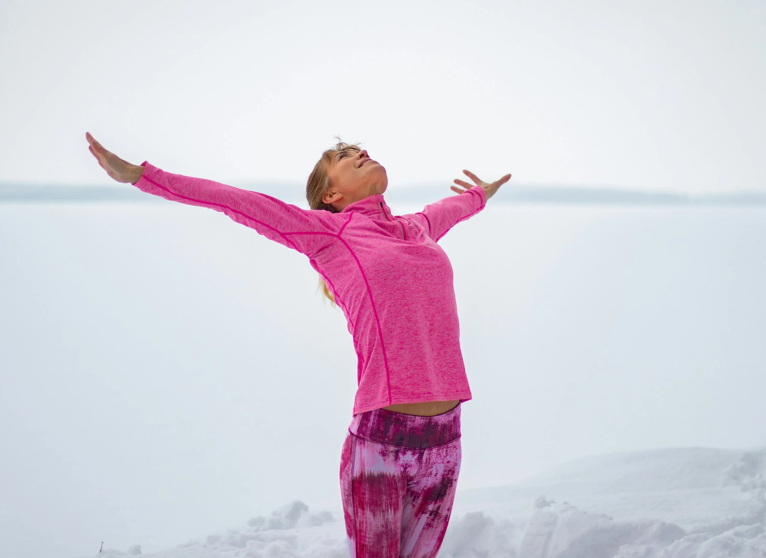 A woman in a pink athletic jacket and pink patterned leggings stretches with her arms outstretched in a snowy landscape.