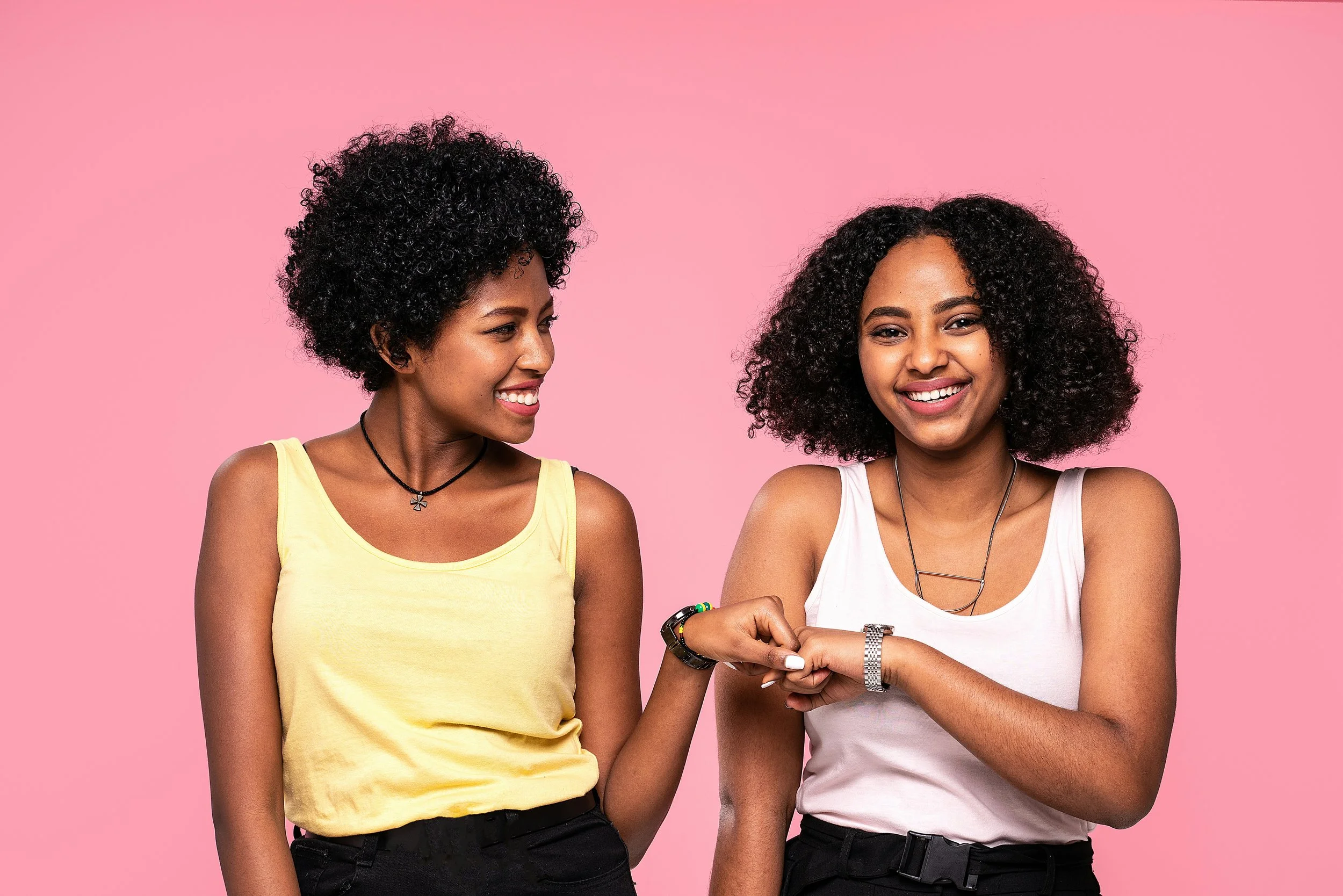 Two women with curly hair smiling and looking at each other against a pink background.