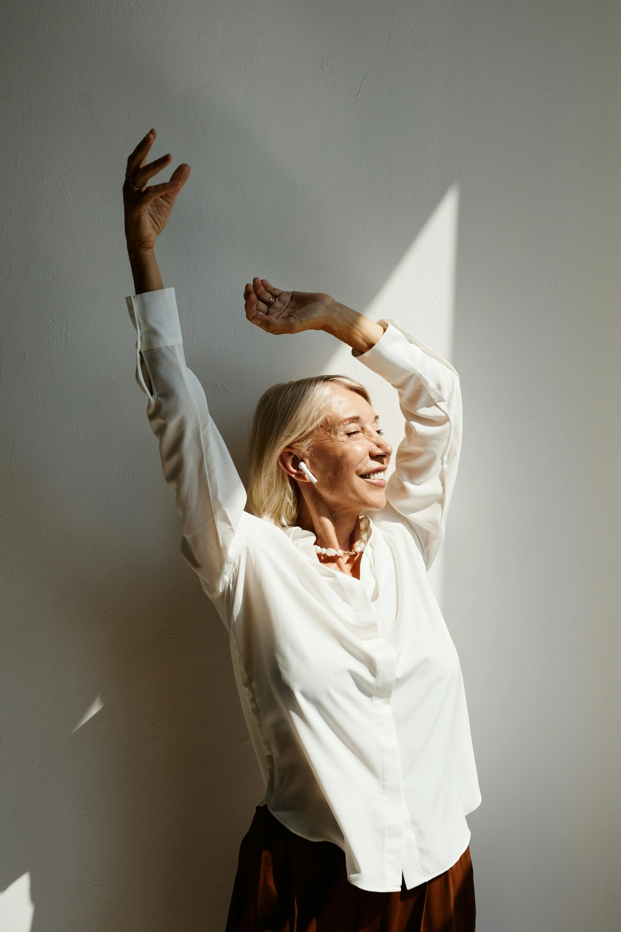 A smiling elderly woman with blonde hair, wearing a white blouse and brown skirt, dancing with her eyes closed and arms raised, standing against a plain wall with sunlight casting shadows.