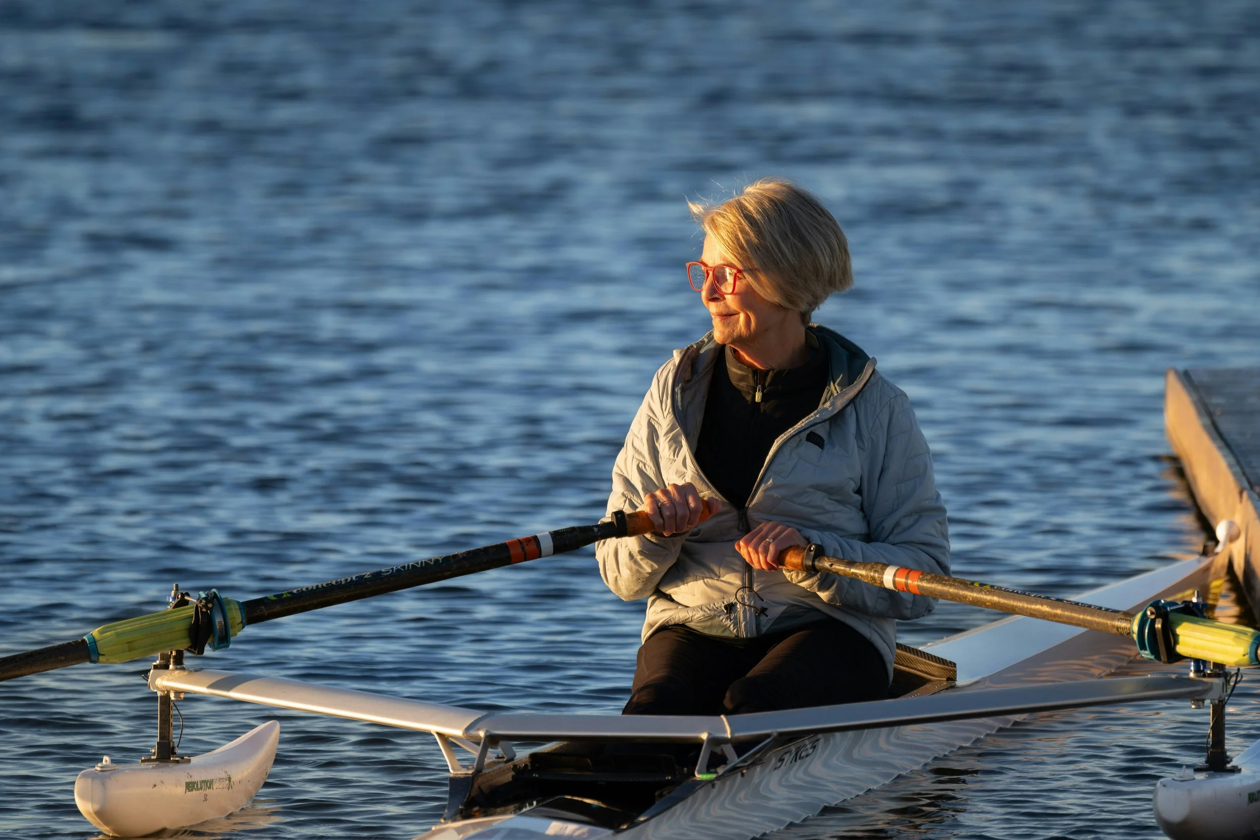 A woman with short blonde hair, wearing red glasses and a light gray jacket, is sitting in a small boat on a body of water, holding oars and smiling while enjoying a sunny day.