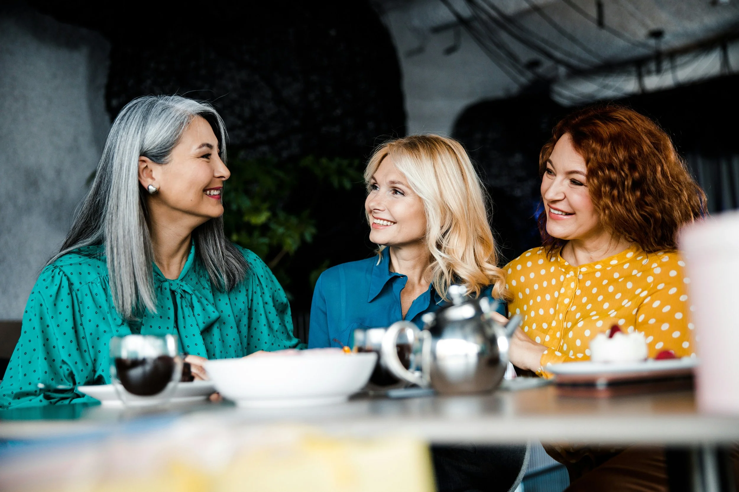 Three women smiling and talking at a table with food and drinks.