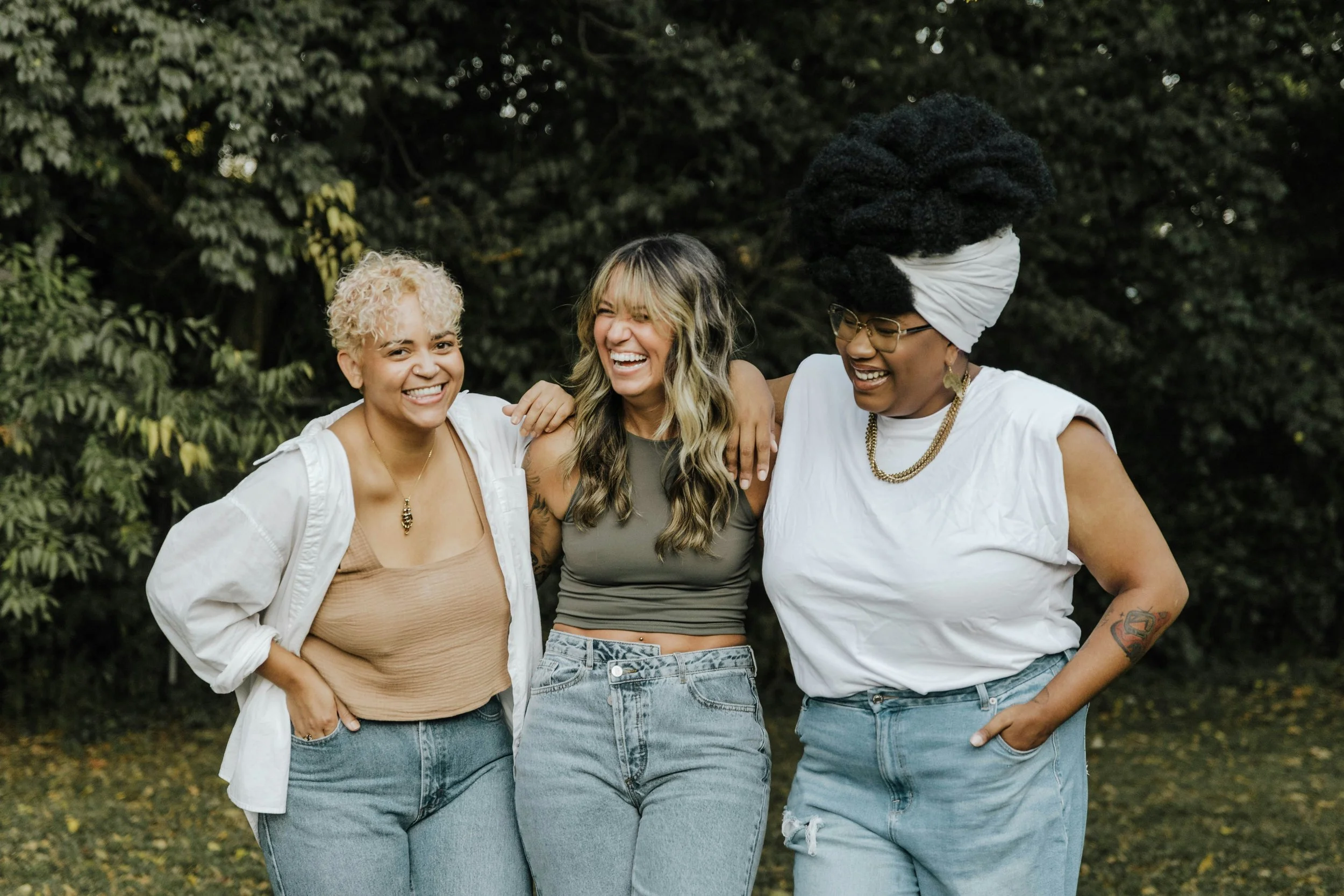 Three women laughing and smiling, standing together outdoors with trees in the background.