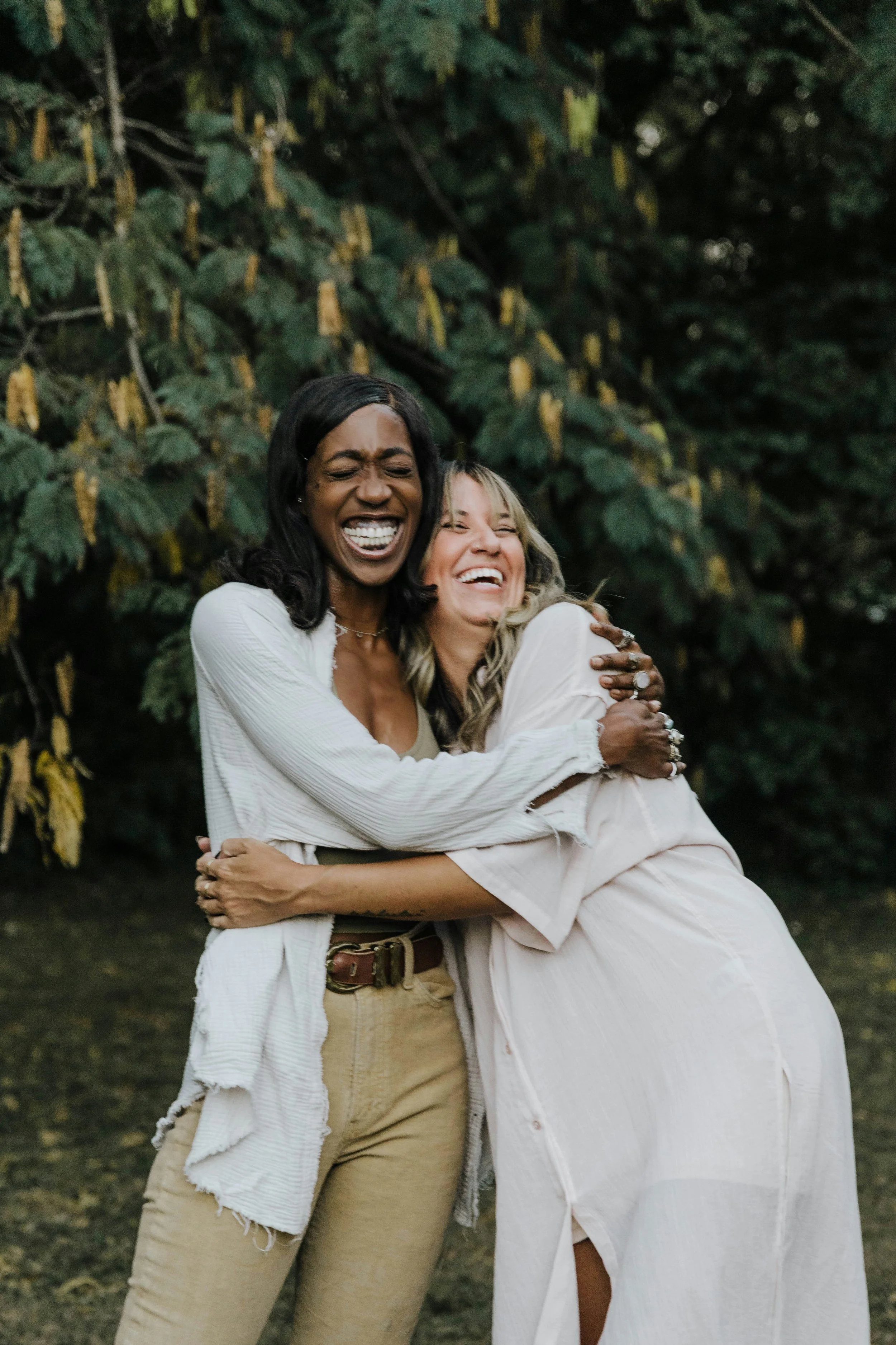 Two women are hugging and smiling outdoors in front of green leafy bushes.